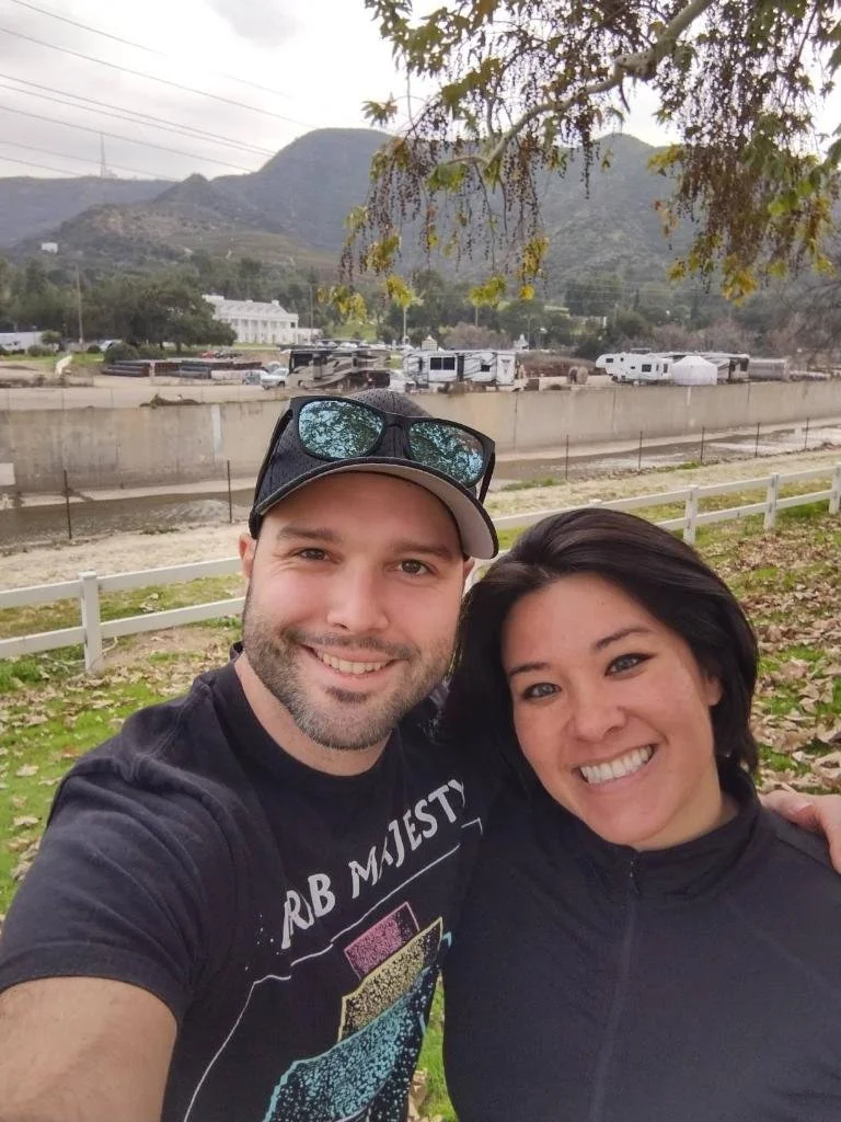 A smiling couple taking a selfie outdoors near a creek, hills, and trees with mountains in the background.