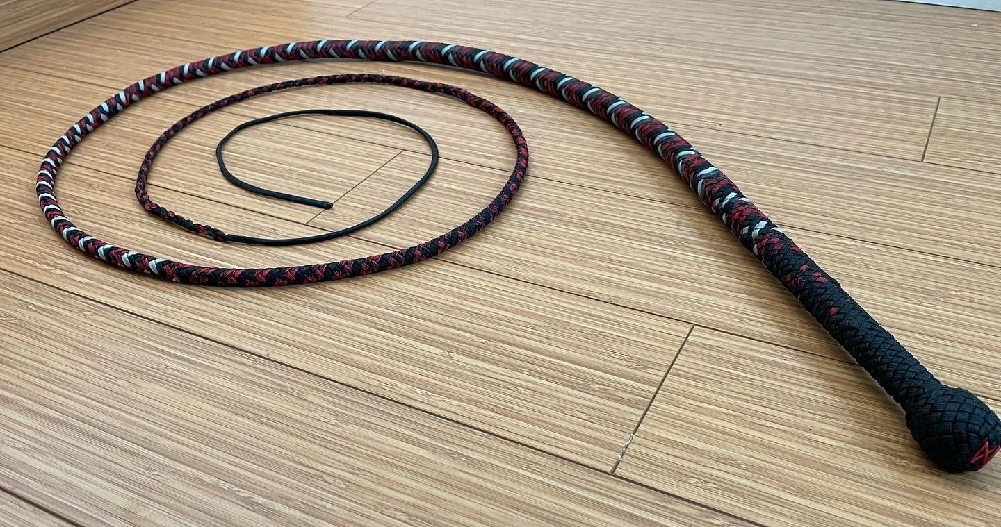 A stripped-braid riding crop with a black handle, a black loop, and a flexible shaft, placed on a wooden floor.