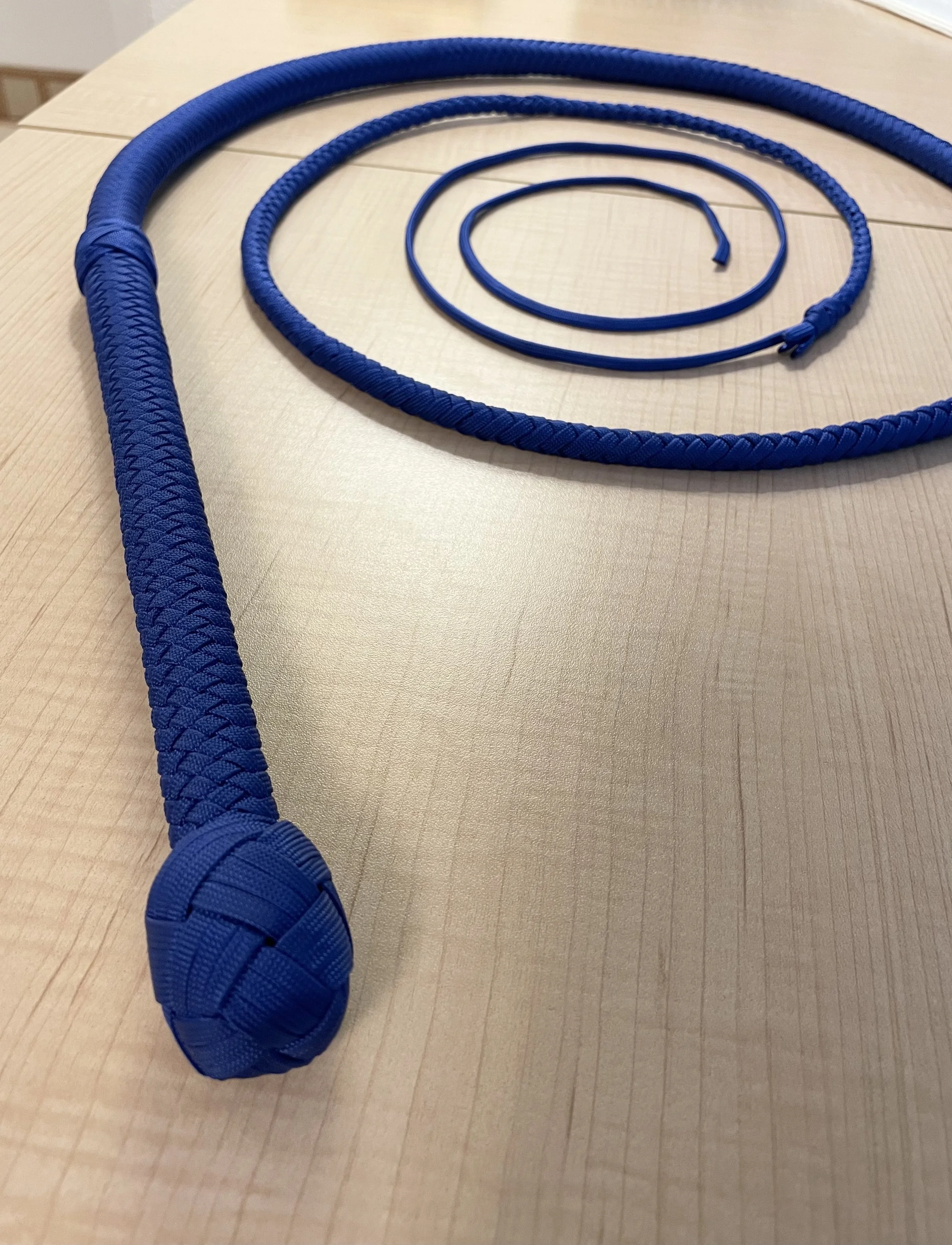 A blue jump rope with a braided handle and a thin, flat cord, laid out on a light wooden surface.
