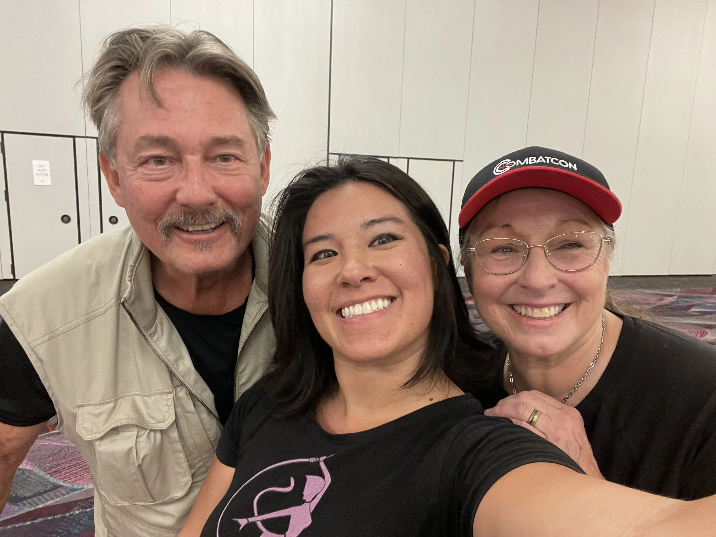 Three smiling people taking a selfie together in an indoor setting with plain walls and a patterned carpet.