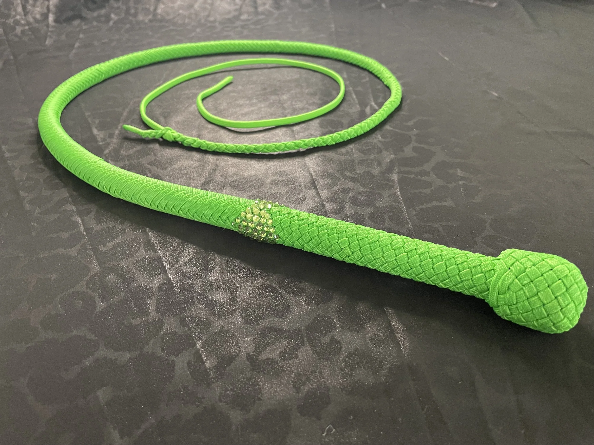 Bright green flexible whip with a braided handle and a small decorative cluster of beads near the handle, lying on a dark surface.