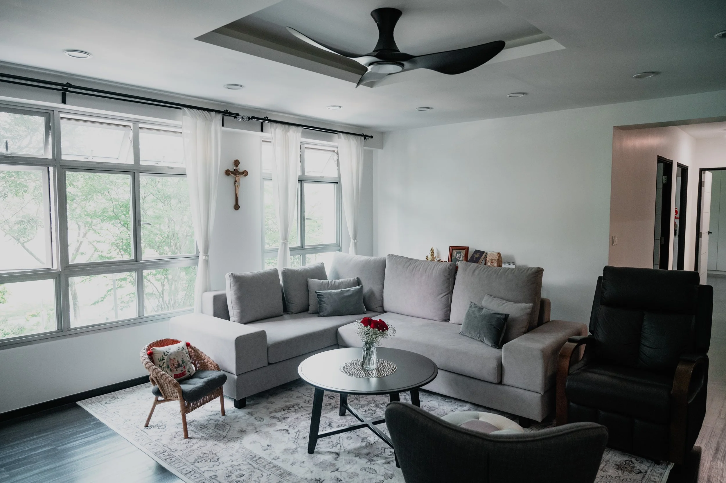 Living room with large gray L-shaped sofa, black armchair, round coffee table with a vase of red flowers, window with white curtains, ceiling fan, and religious cross on the wall.