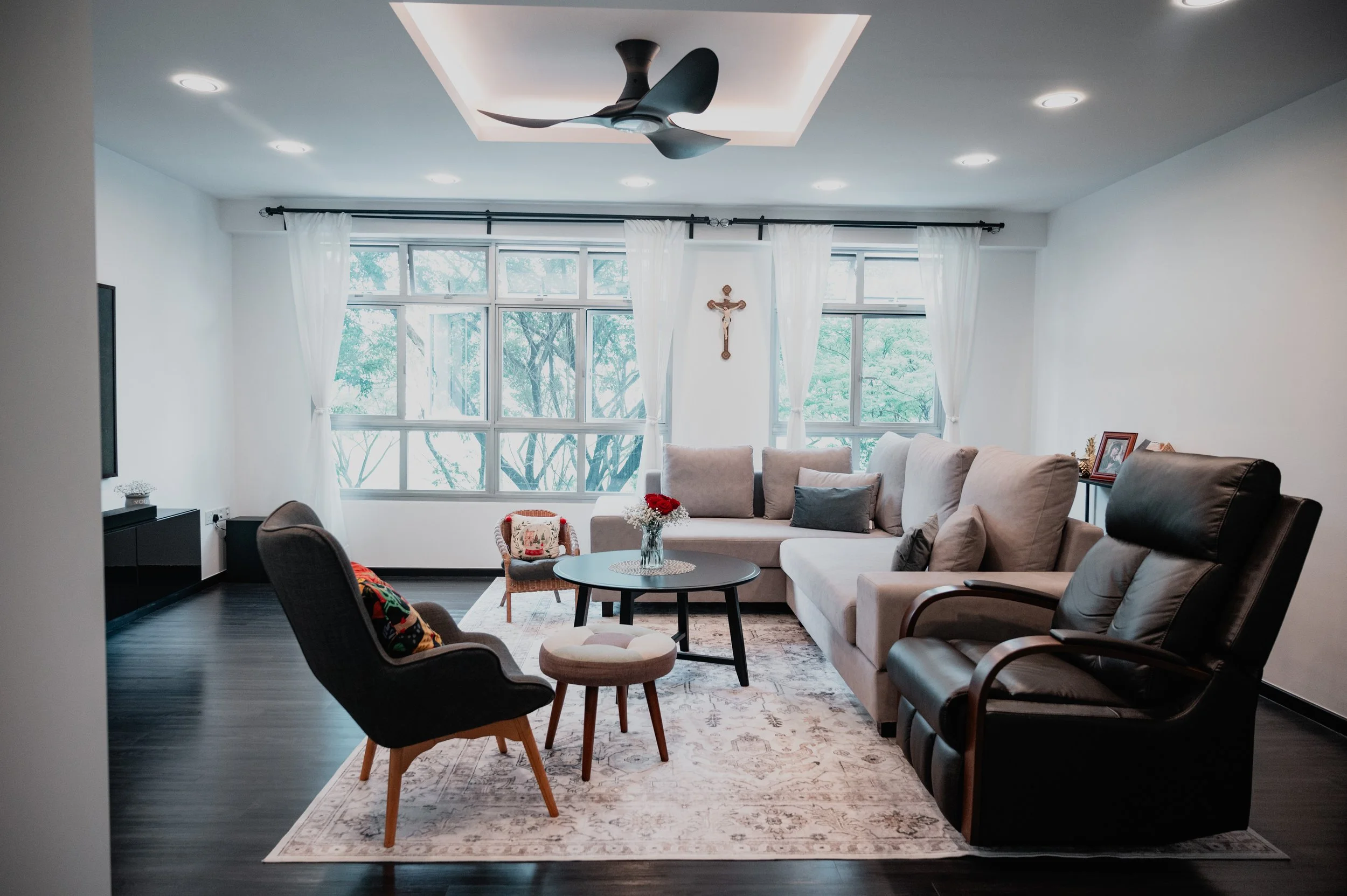 Living room with large windows, white curtains, beige sectional sofa, black leather armchair, coffee table with vase of red flowers, and crucifix on the wall.
