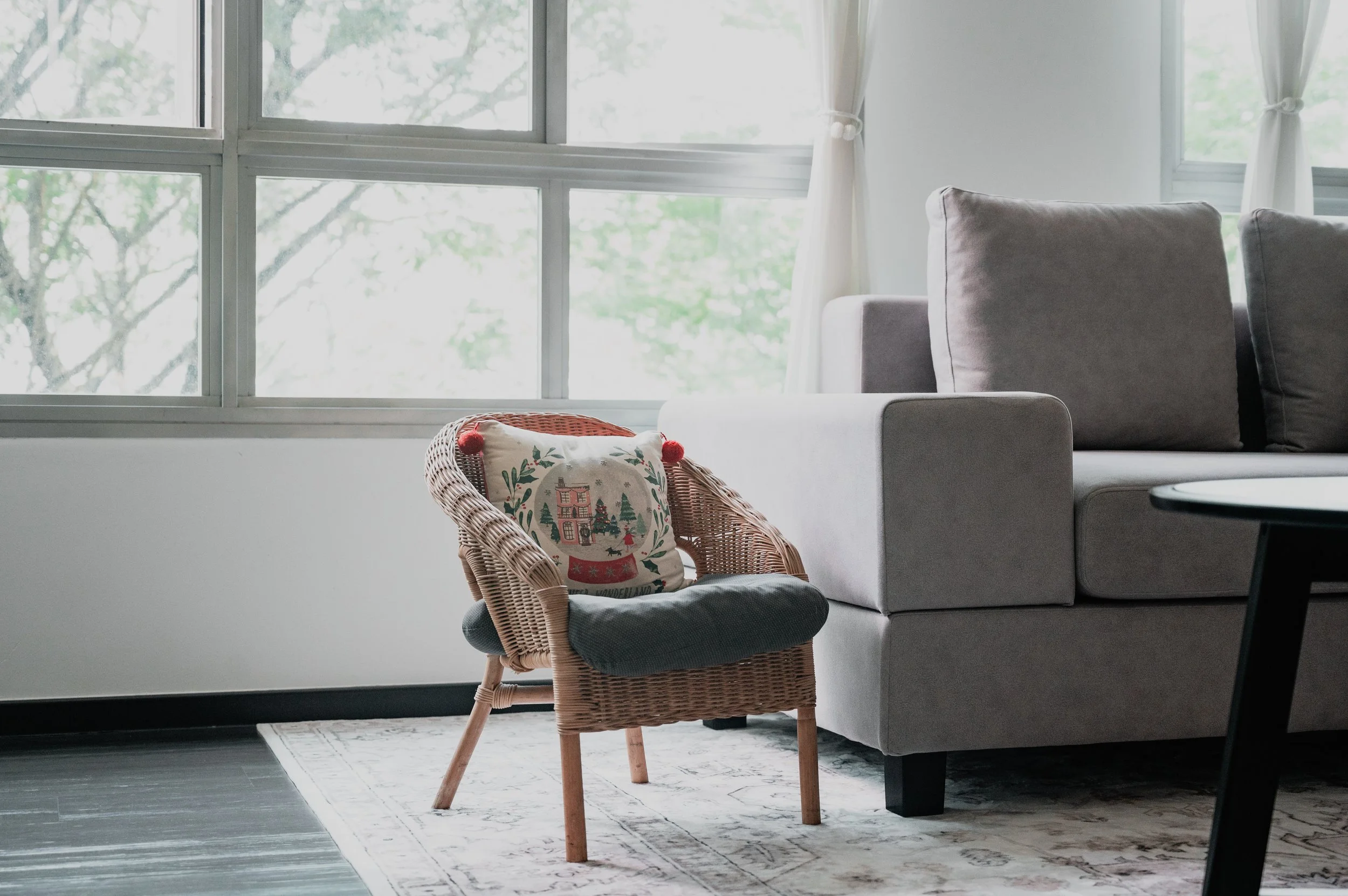 Interior of a living room with large windows, a gray sofa, a wicker chair with a holiday-themed cushion, a patterned rug, and a small round black table.