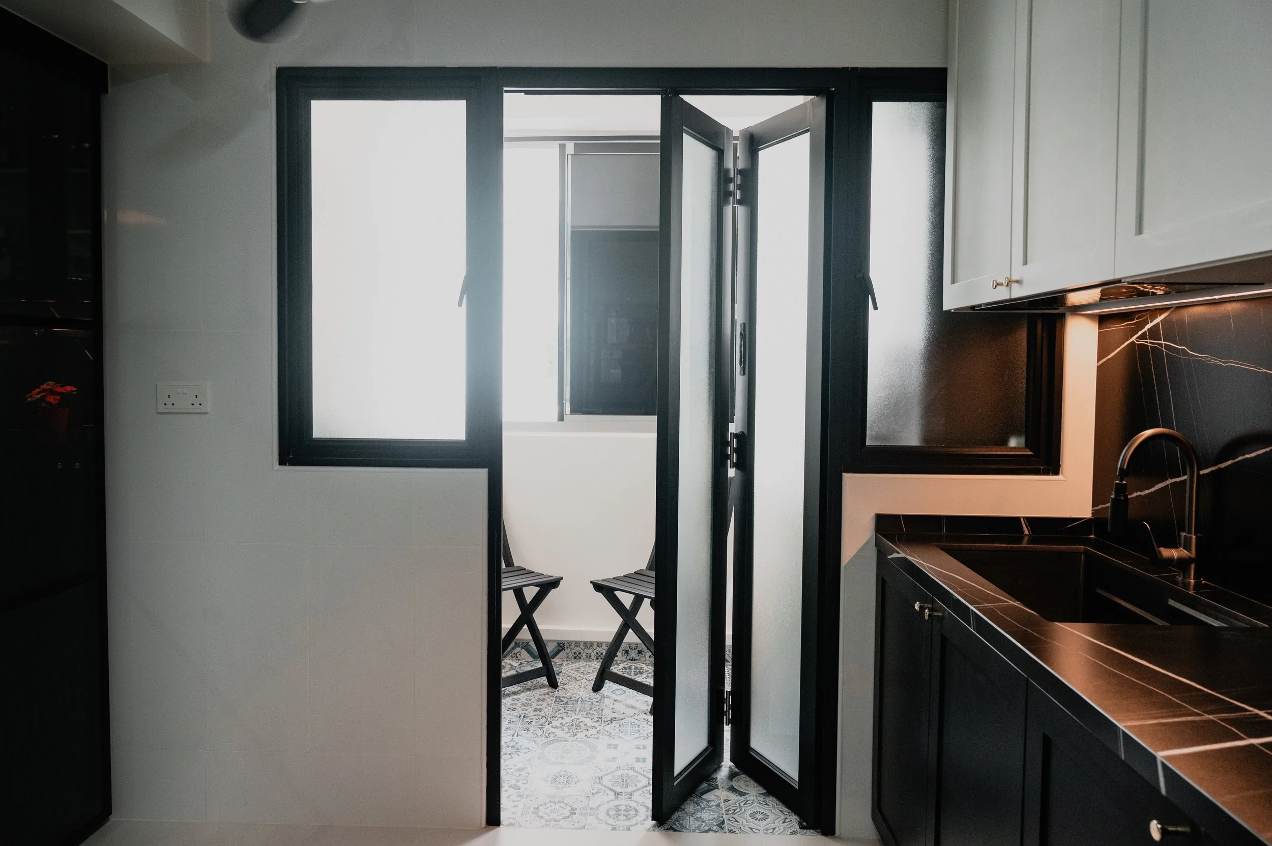 Kitchen area with black countertops, white upper cabinets, and a double window with frosted glass. A small balcony with two black folding chairs is visible through the open glass door.