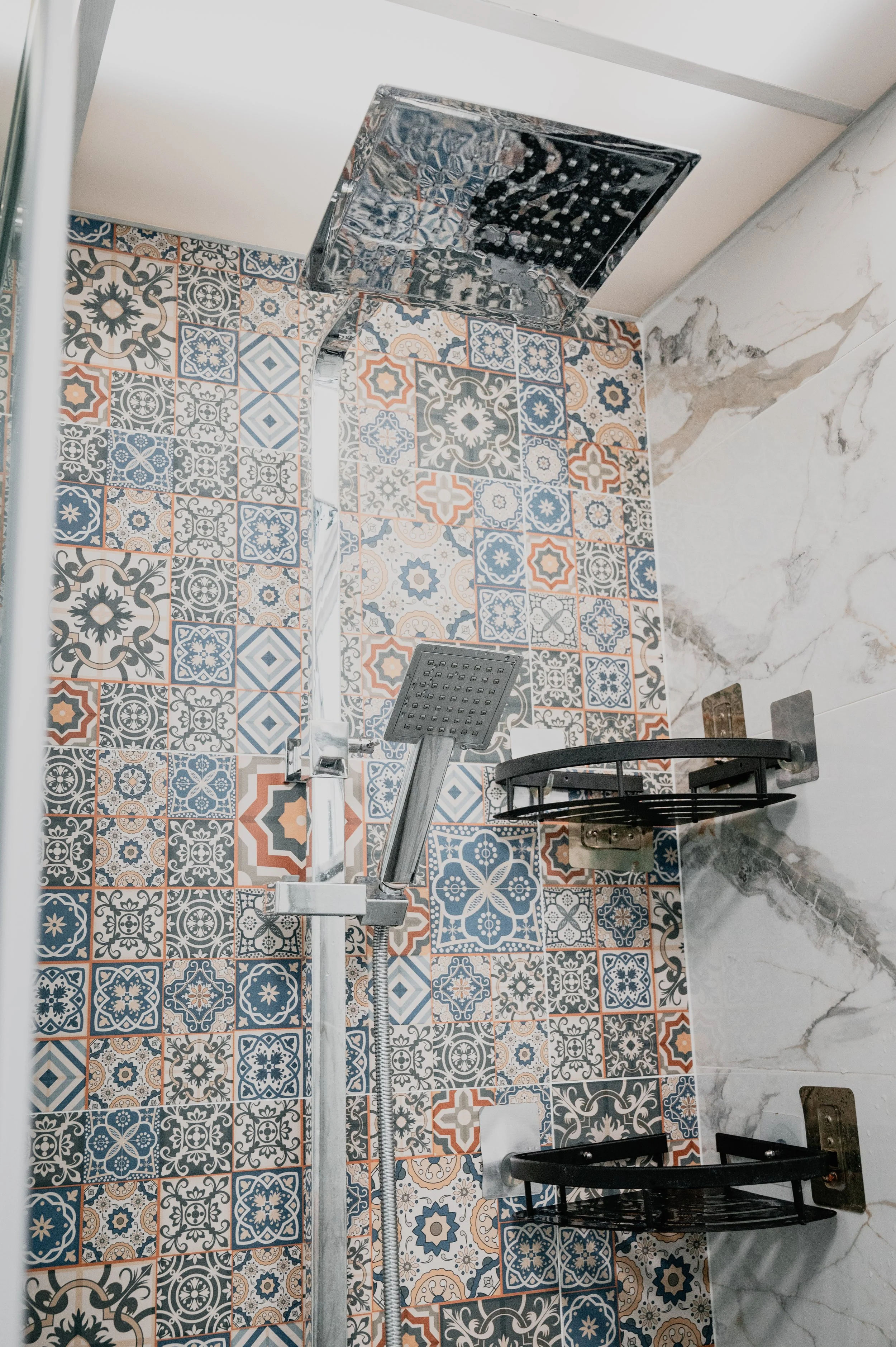 A decorative tiled shower wall with a rain shower head, a smaller handheld shower head, a soap dish, and marble accents.