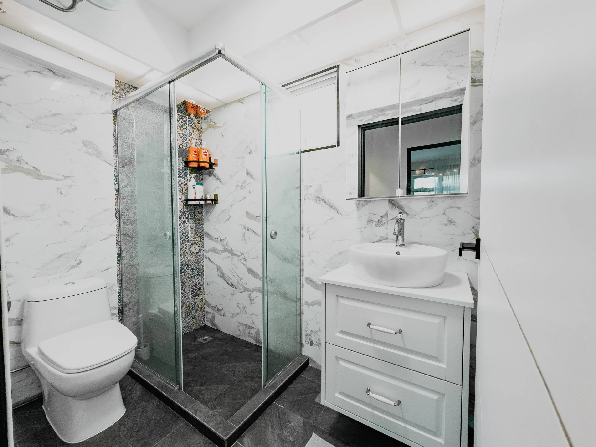 Modern bathroom with white marble walls, a glass shower enclosure with patterned tile accent wall, a white vanity with a vessel sink, a mirror above, and a toilet nearby.