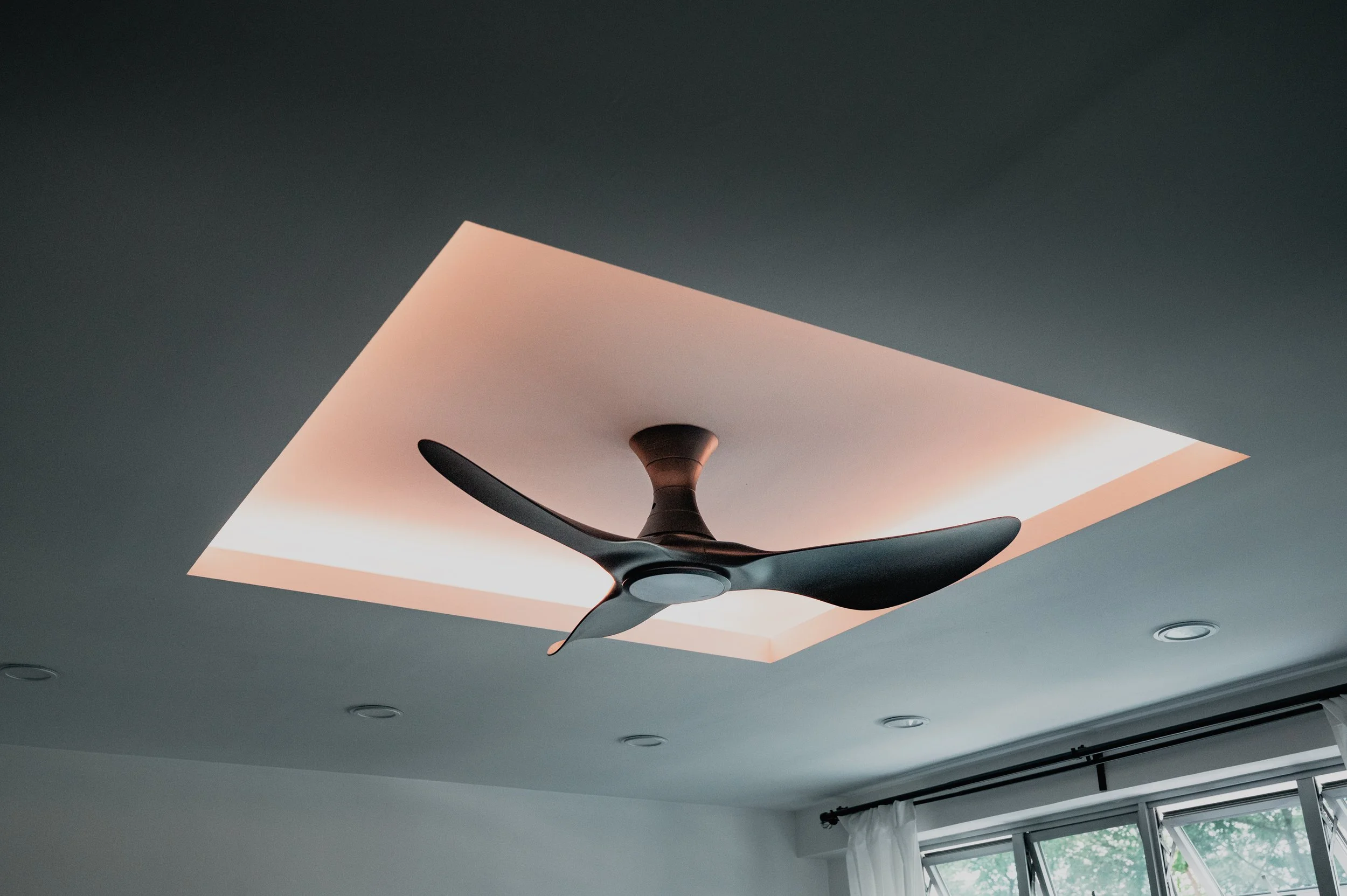 Modern ceiling fan with three blades installed on a ceiling with a rectangular skylight and recessed lighting, beside a window with white curtains.