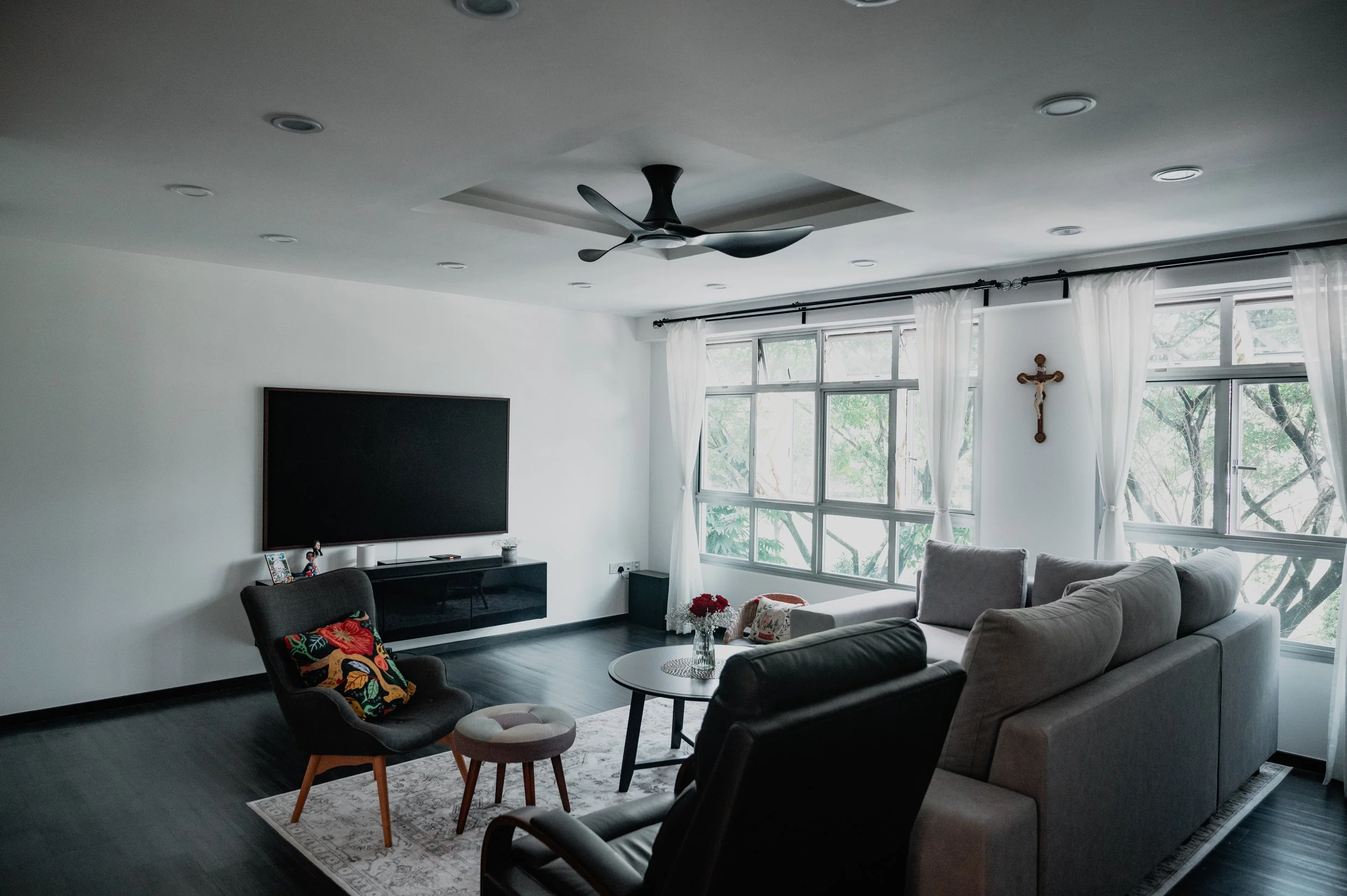 Living room with large windows, white walls, black flooring, a gray sectional sofa, a black armchair with a colorful pillow, a small ottoman, a round coffee table with a vase of red flowers, a wall-mounted TV, a ceiling fan, white curtains, and a cru