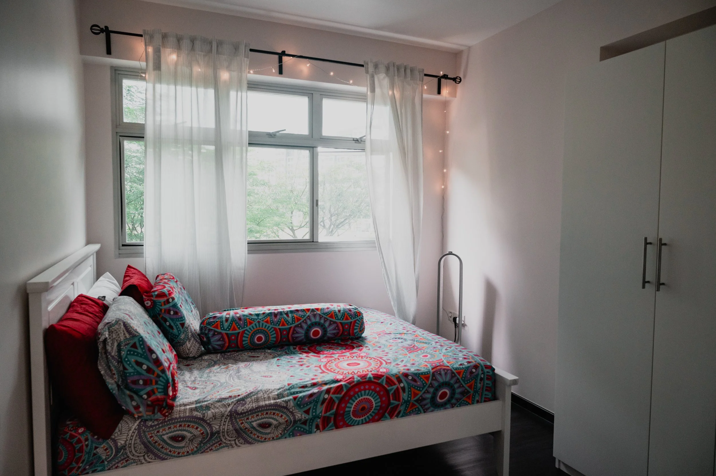 A bedroom with a white bed, colorful patterned bedding, red pillows, sheer white curtains, and a white wardrobe.