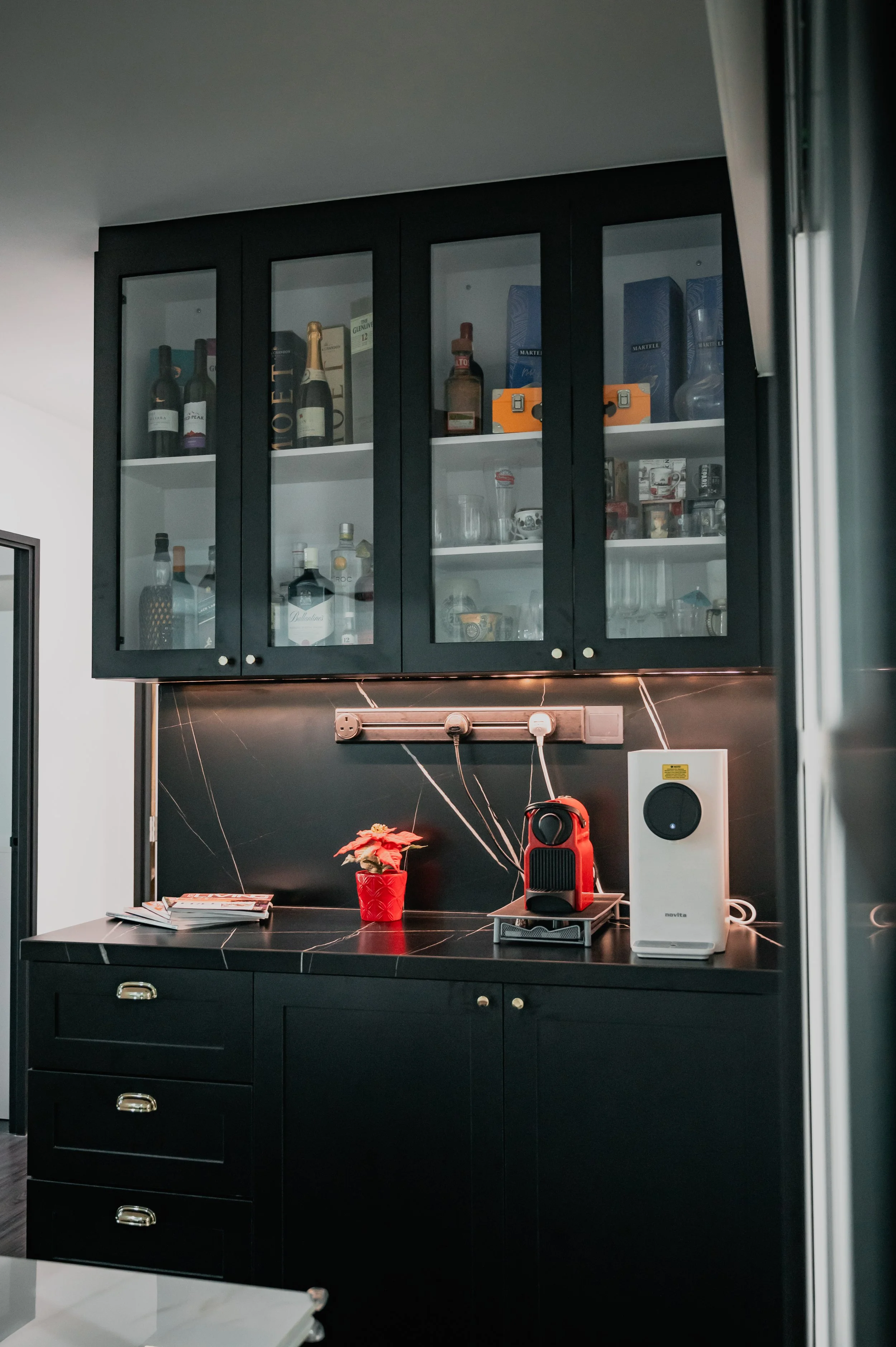 Black kitchen cabinet with glass doors containing bottles and glasses, black countertop with small flower pot, red coffee machine, and white air purifier.