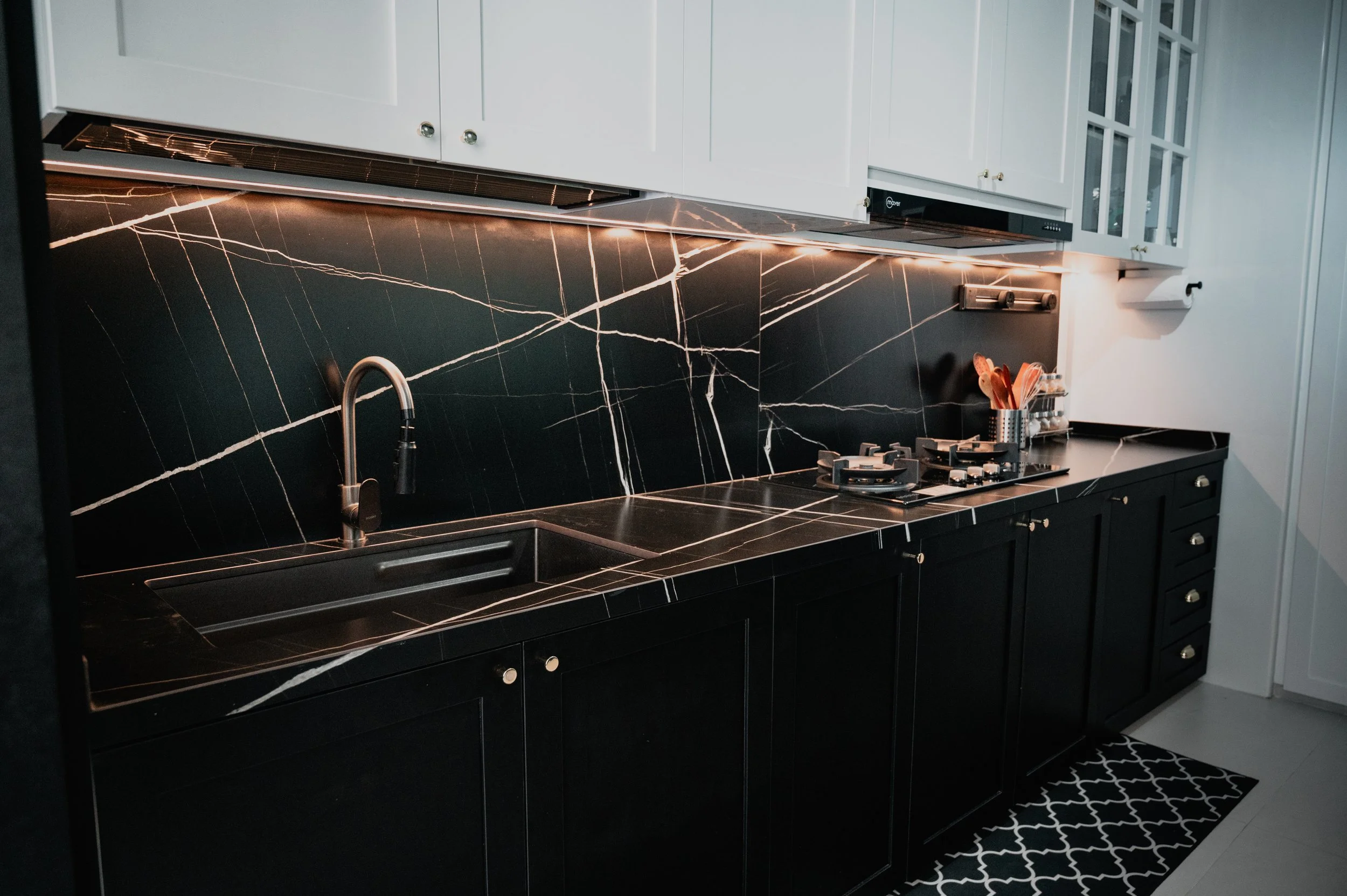 Modern kitchen with black marble countertops and backsplash, white upper cabinets, and black lower cabinets. Includes a sink, stove, and utensils in a holder on the counter.