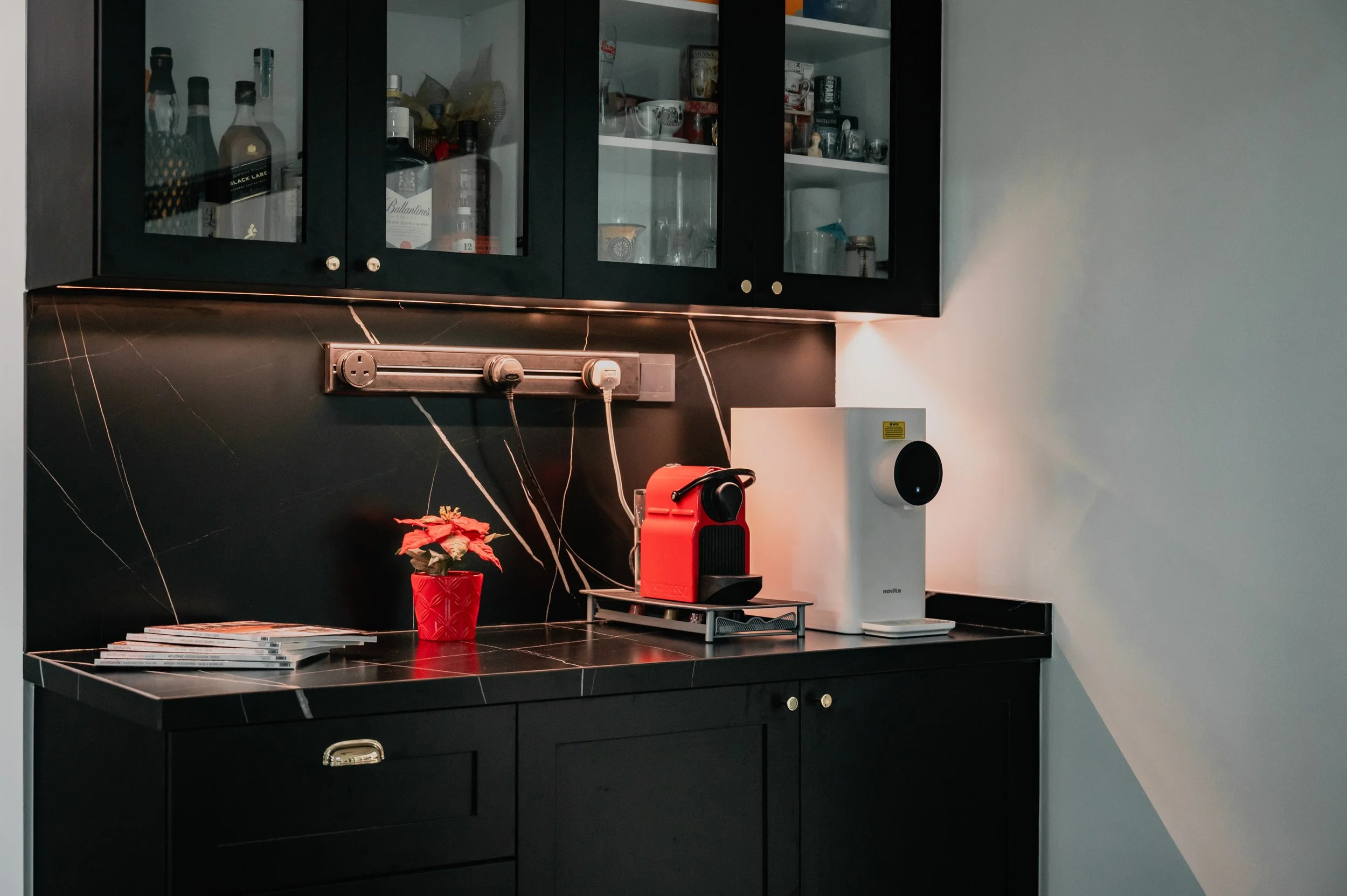 A black kitchen counter with a red coffee machine, a white speaker, a small poinsettia plant in a red pot, and magazines. Black cabinets with glass doors are above, with various bottles and kitchen items inside.