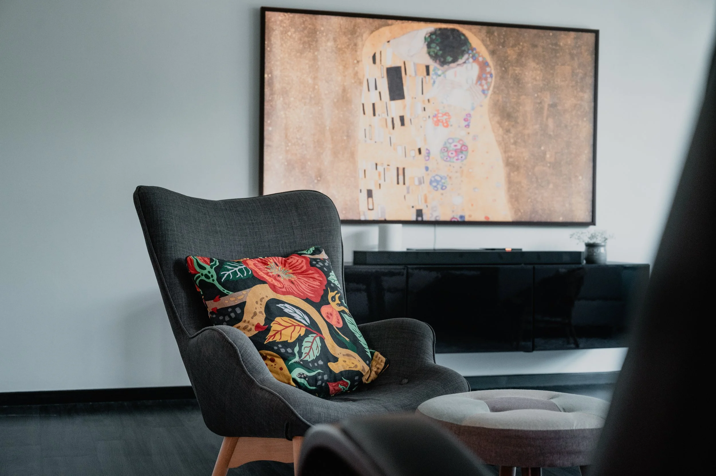 A modern living room corner with a gray armchair, a colorful floral cushion, a round ottoman, a large flat-screen television, and a framed artwork on the wall, with a black TV stand and a small potted plant.