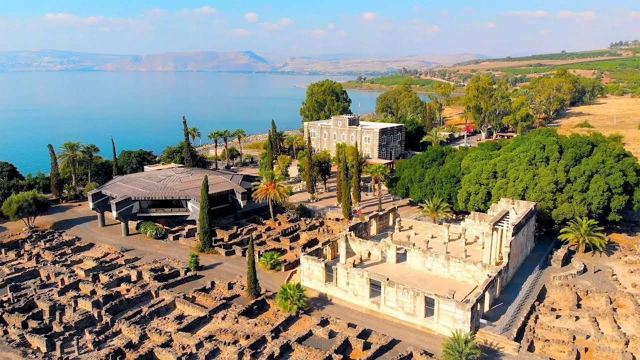 Aerial view of ancient ruins with a modern building and lush greenery near a large body of water, with hills in the background.