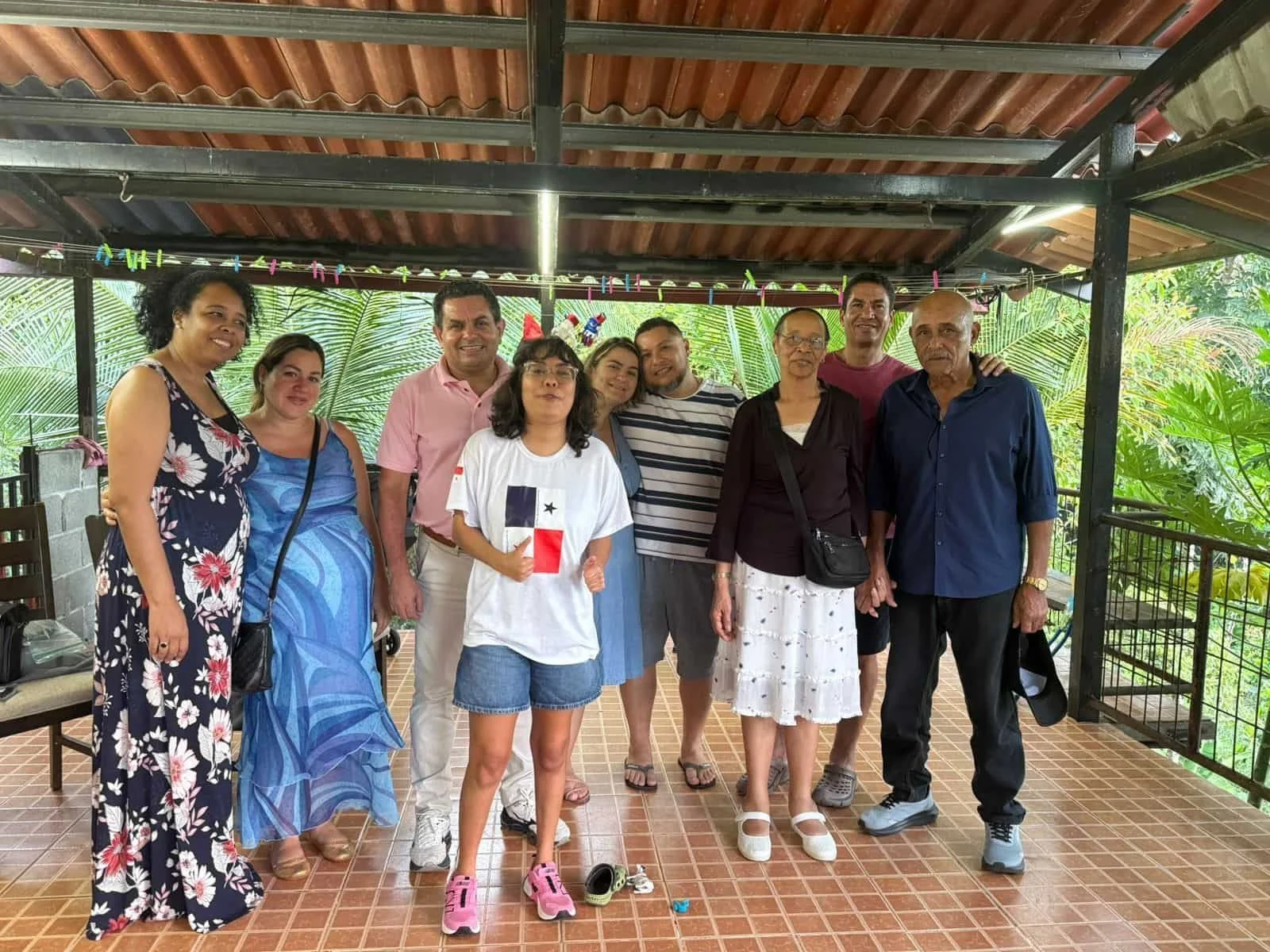 A group of eleven people standing together on a tiled porch with palm trees in the background, some smiling and some making gestures.