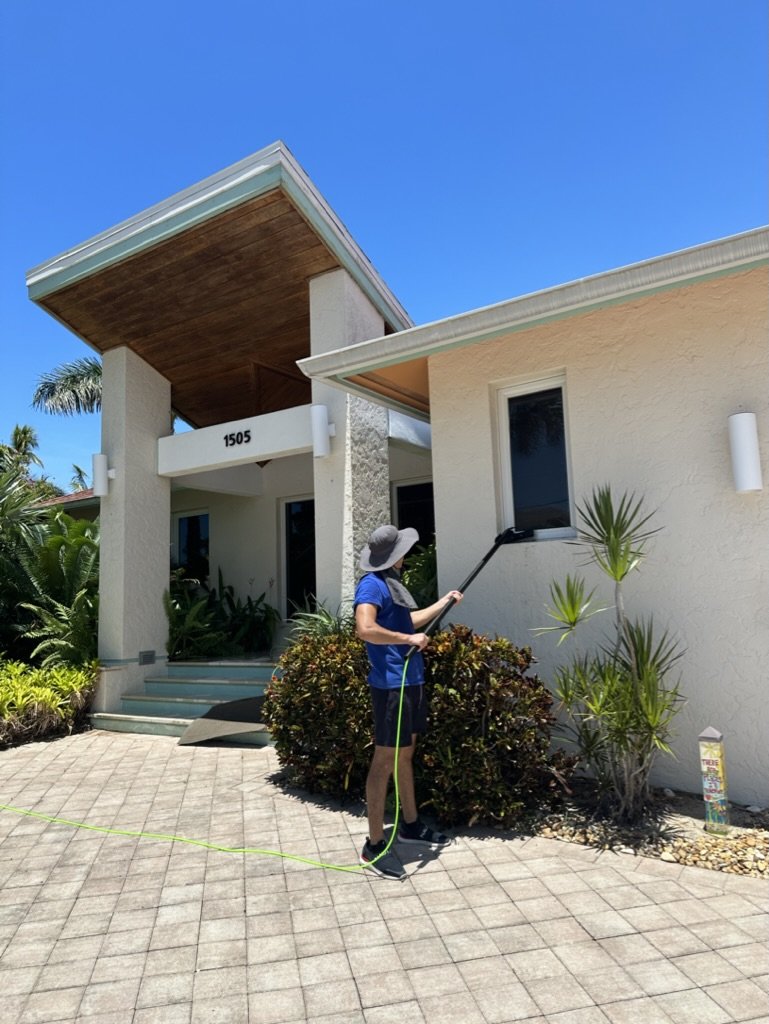 Person cleaning a window of a modern house with a tall entrance under a clear blue sky, surrounded by greenery and a paved driveway.