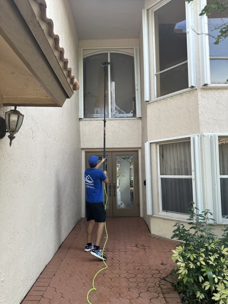 Person cleaning second-story window with a long pole, wearing a blue shirt and cap, standing on a red brick walkway.