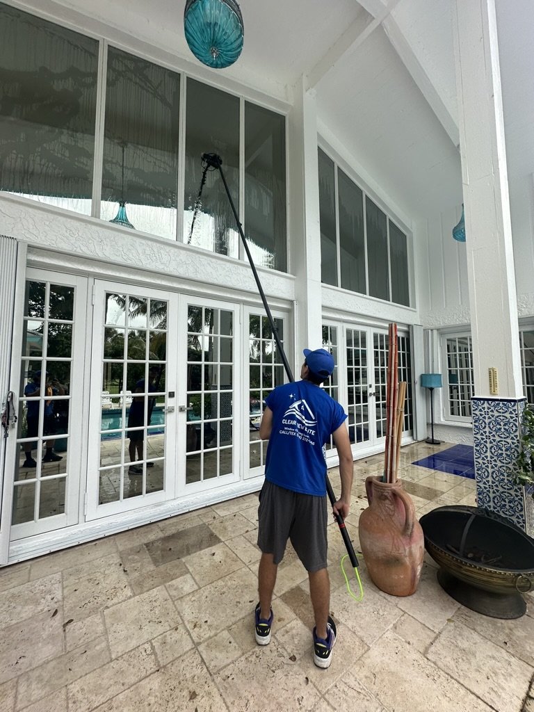 Person cleaning large windows with a long pole on a patio with tile flooring and decorative items.