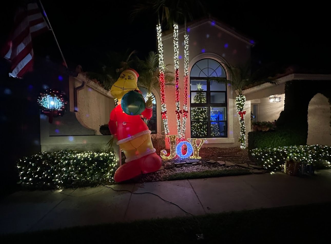 House decorated with Christmas lights, inflatable Grinch in Santa outfit, JOY sign, and Christmas tree visible through window.