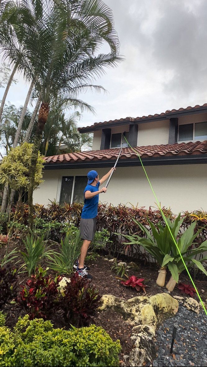 Man in a blue shirt and cap using a telescopic pole to clean the second-floor window of a house with a tiled roof, surrounded by tropical plants and shrubs.