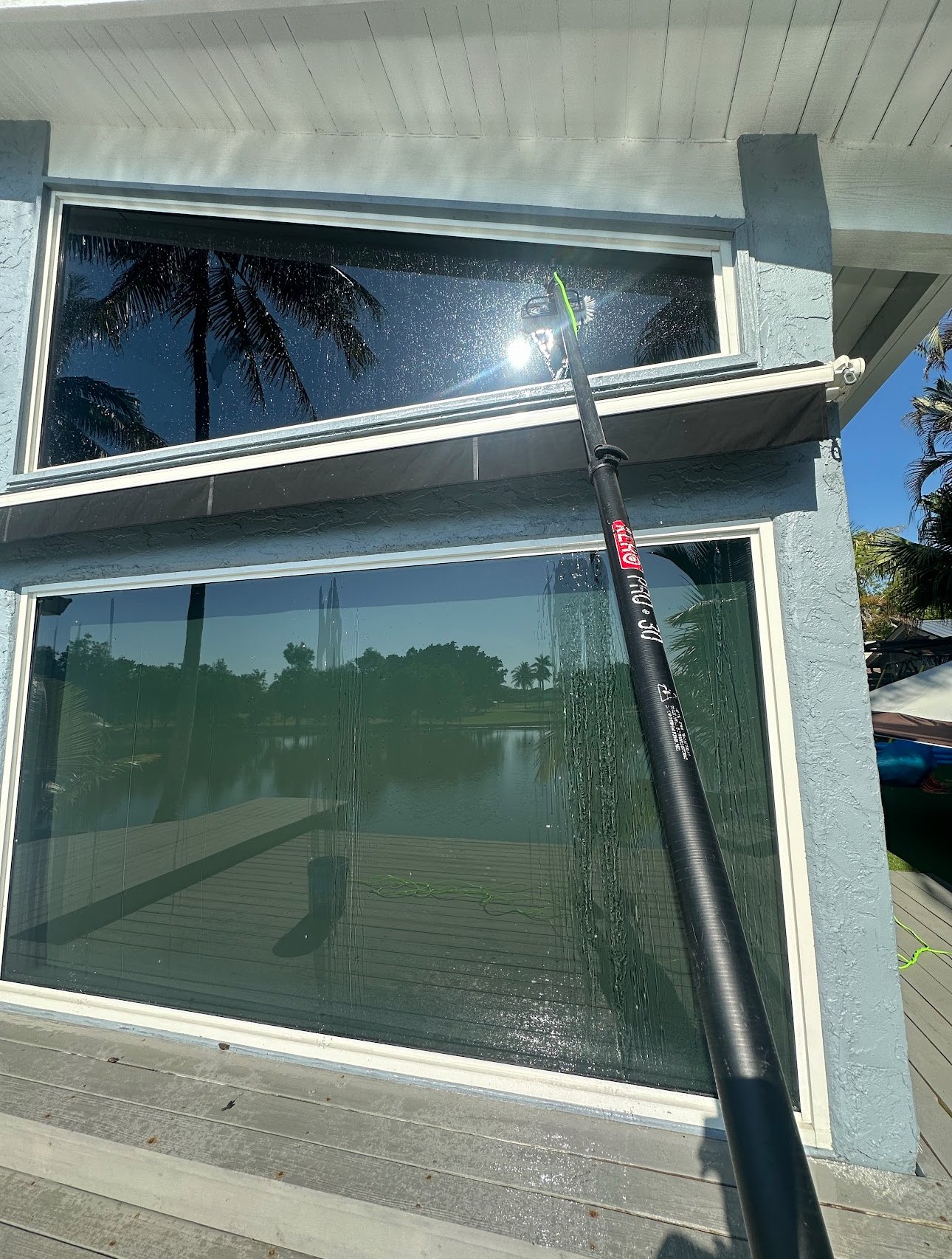A large window being cleaned with a long-handled brush on a sunny day, reflecting palm trees and a body of water.