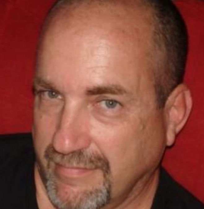 Close-up of a middle-aged man with short dark hair, a goatee, and blue eyes, looking at the camera with a slight smile against a red background.
