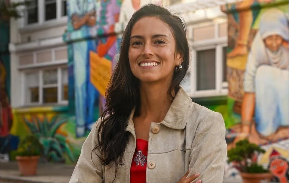 A woman with long dark hair stands outdoors, smiling with arms crossed, in front of a colorful mural.
