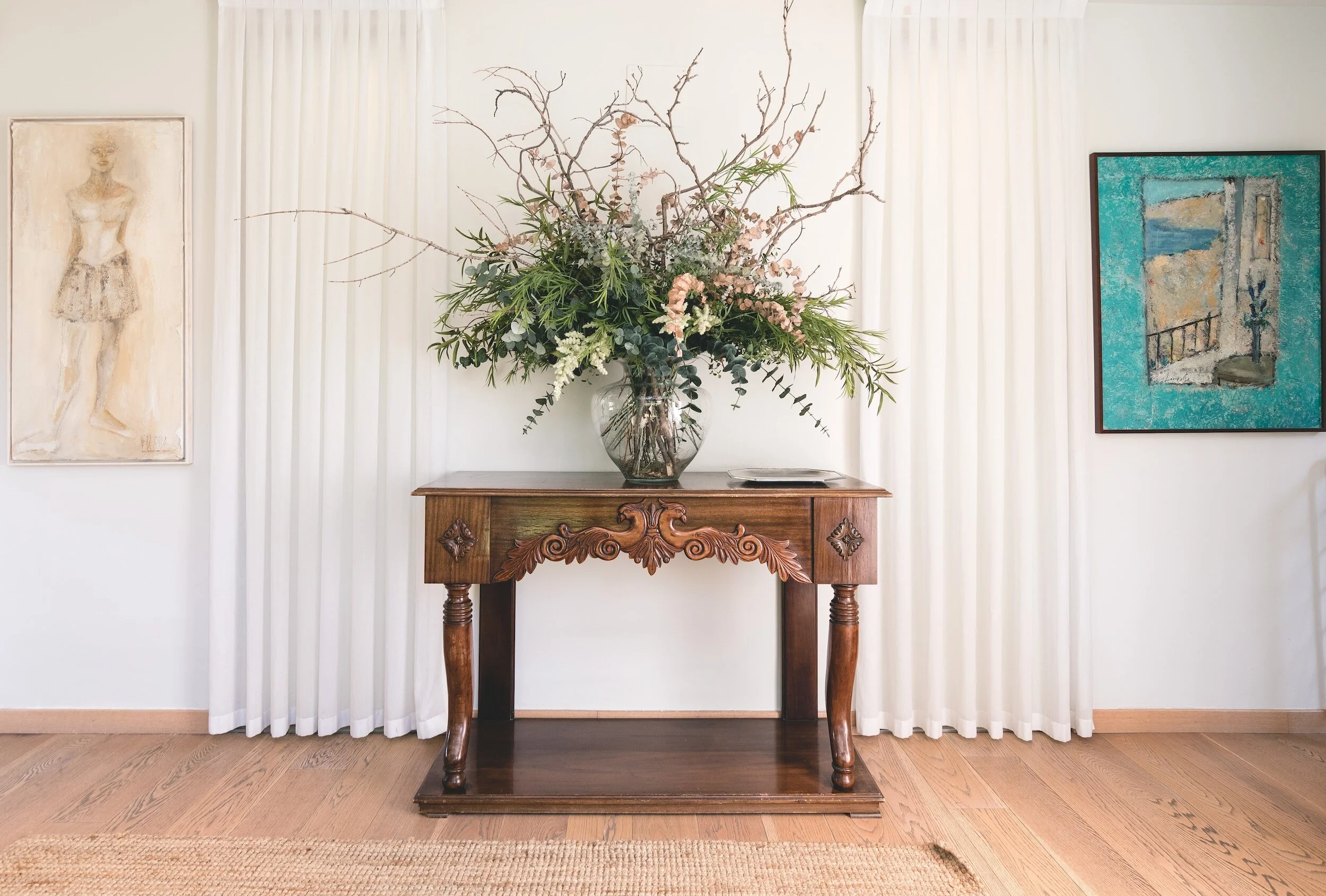 A decorated wooden console table with a large floral arrangement in a glass vase, flanked by two framed paintings on a white wall with curtains in the background.
