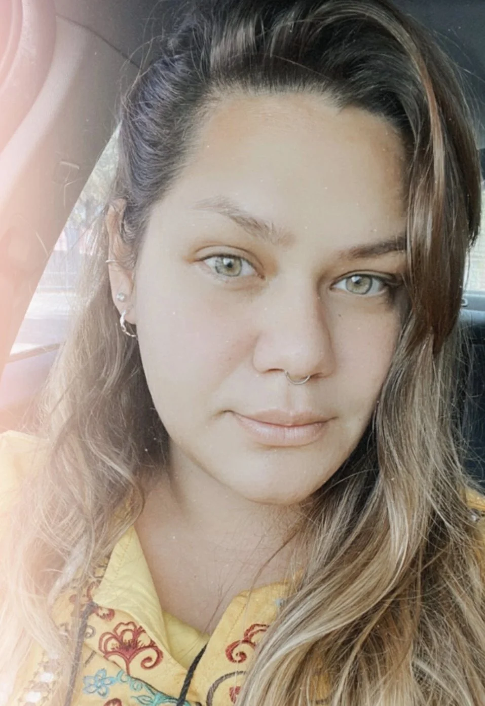 Close-up of a young woman with long, wavy hair, blue eyes, and a septum piercing, wearing a yellow top with red and blue embroidery, sitting inside a vehicle.