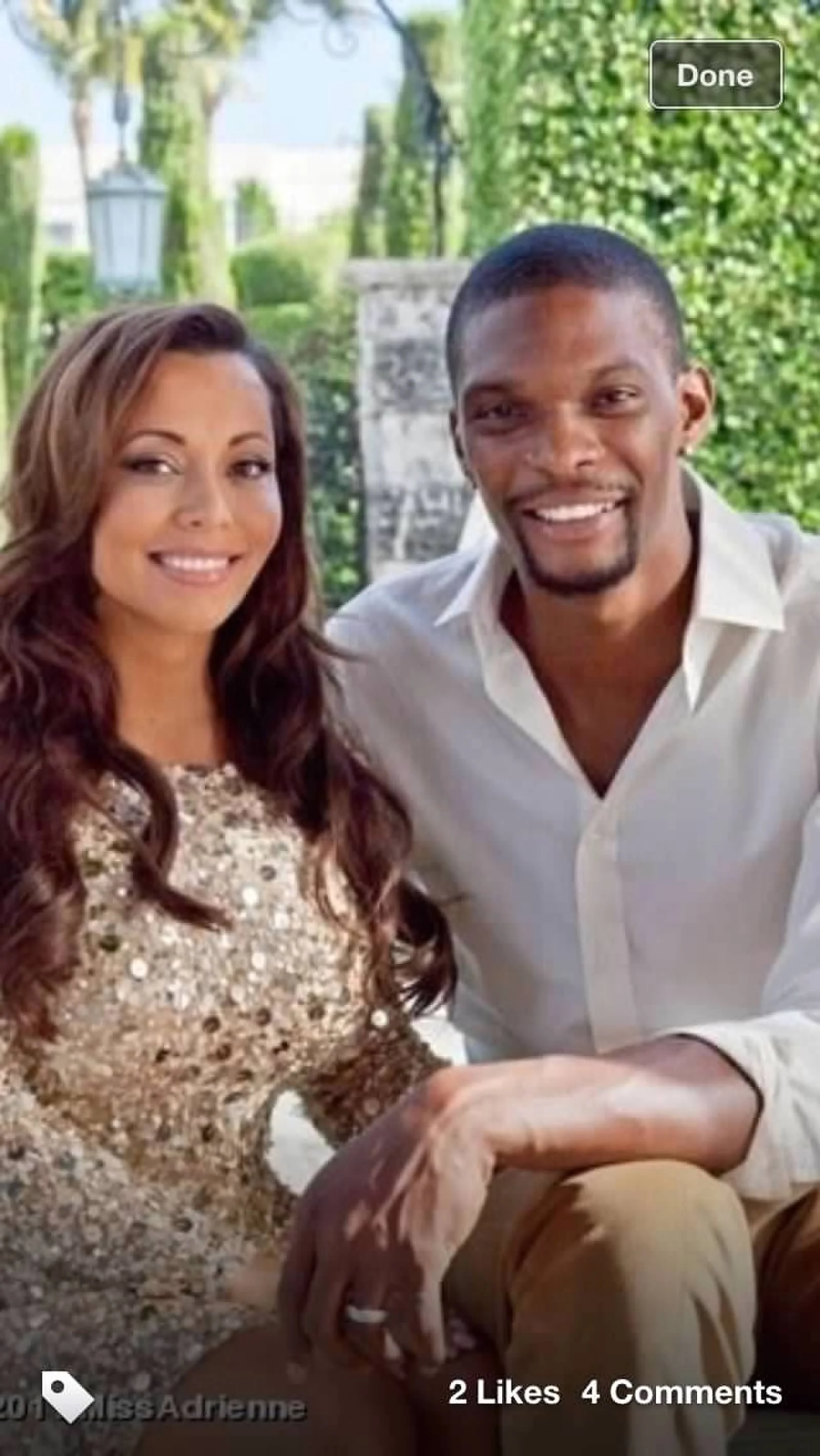 A smiling woman with long wavy brown hair wearing a gold sequined dress, and a smiling man with short black hair and a goatee wearing a white shirt, sitting outdoors with green trees and a park-like setting in the background.