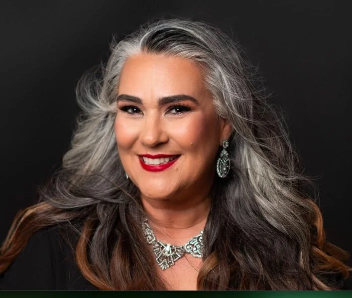 Portrait of a smiling older woman with long, wavy gray and brown hair wearing jewelry and a black top against a dark background.