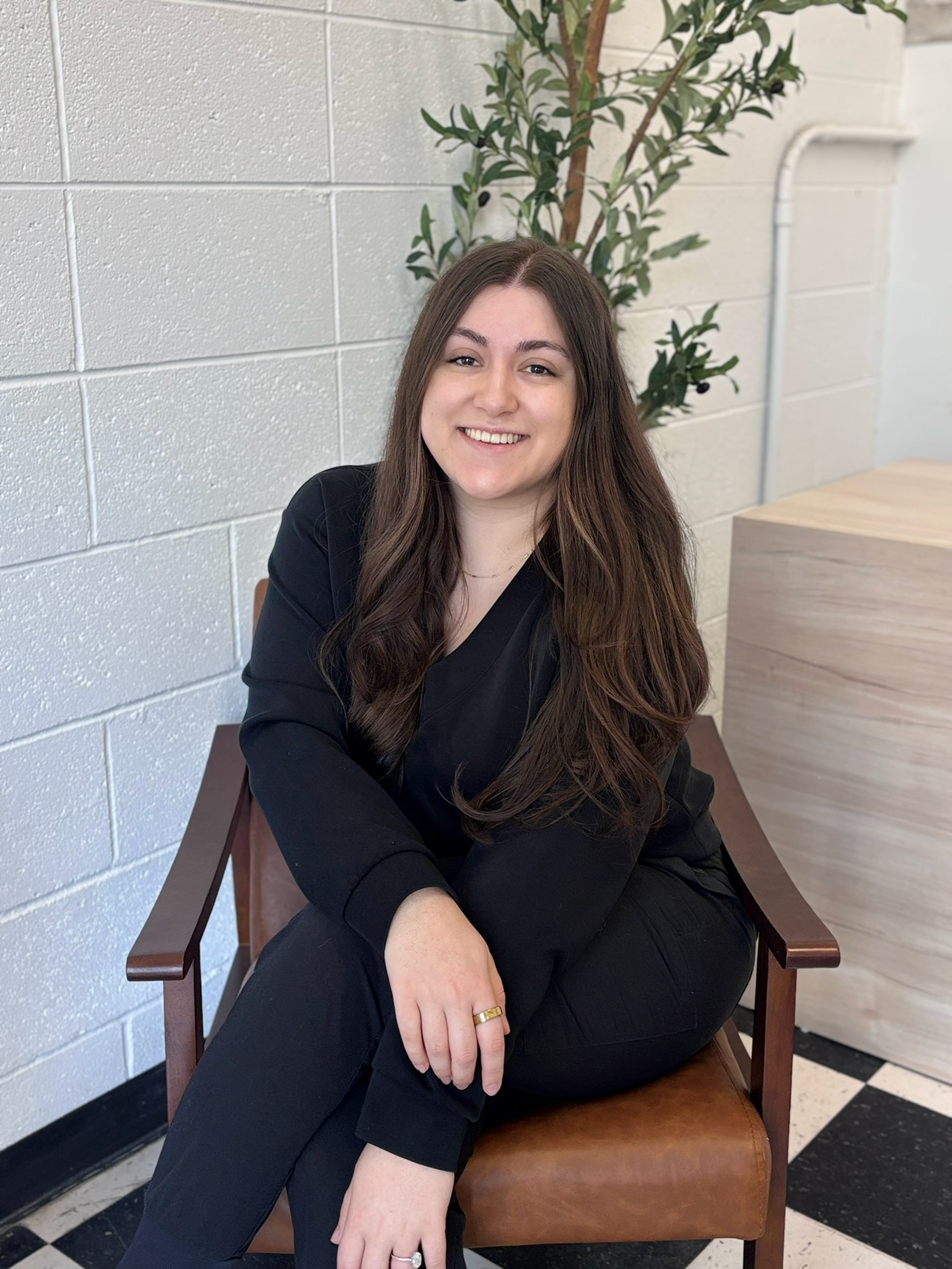 A young woman with long brown hair, smiling, wearing a black blazer and pants, sitting on a wooden chair in a room with white cinderblock walls and a green plant behind her.