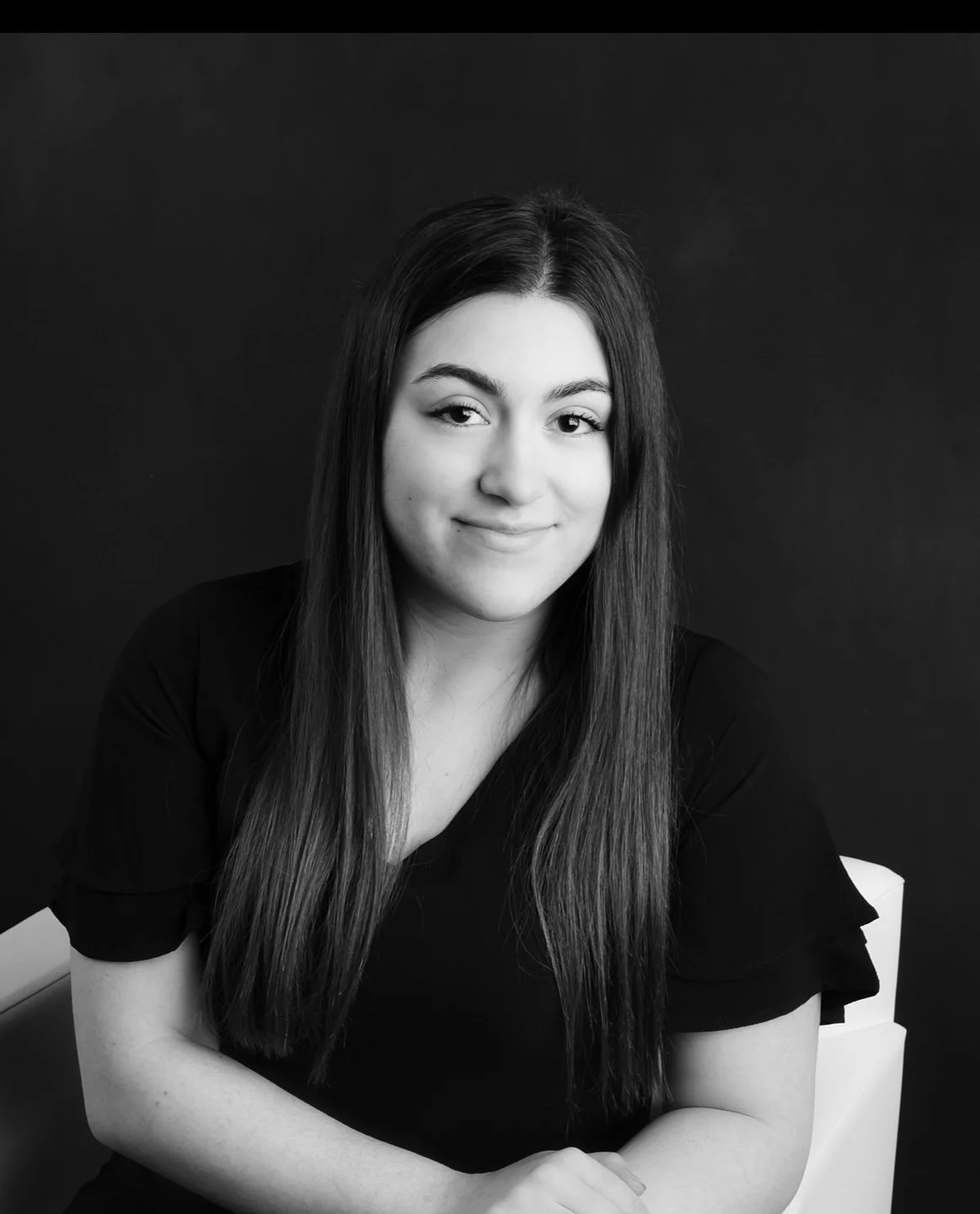 Black and white portrait of a young woman with long hair, smiling gently, sitting on a white chair against a dark background.