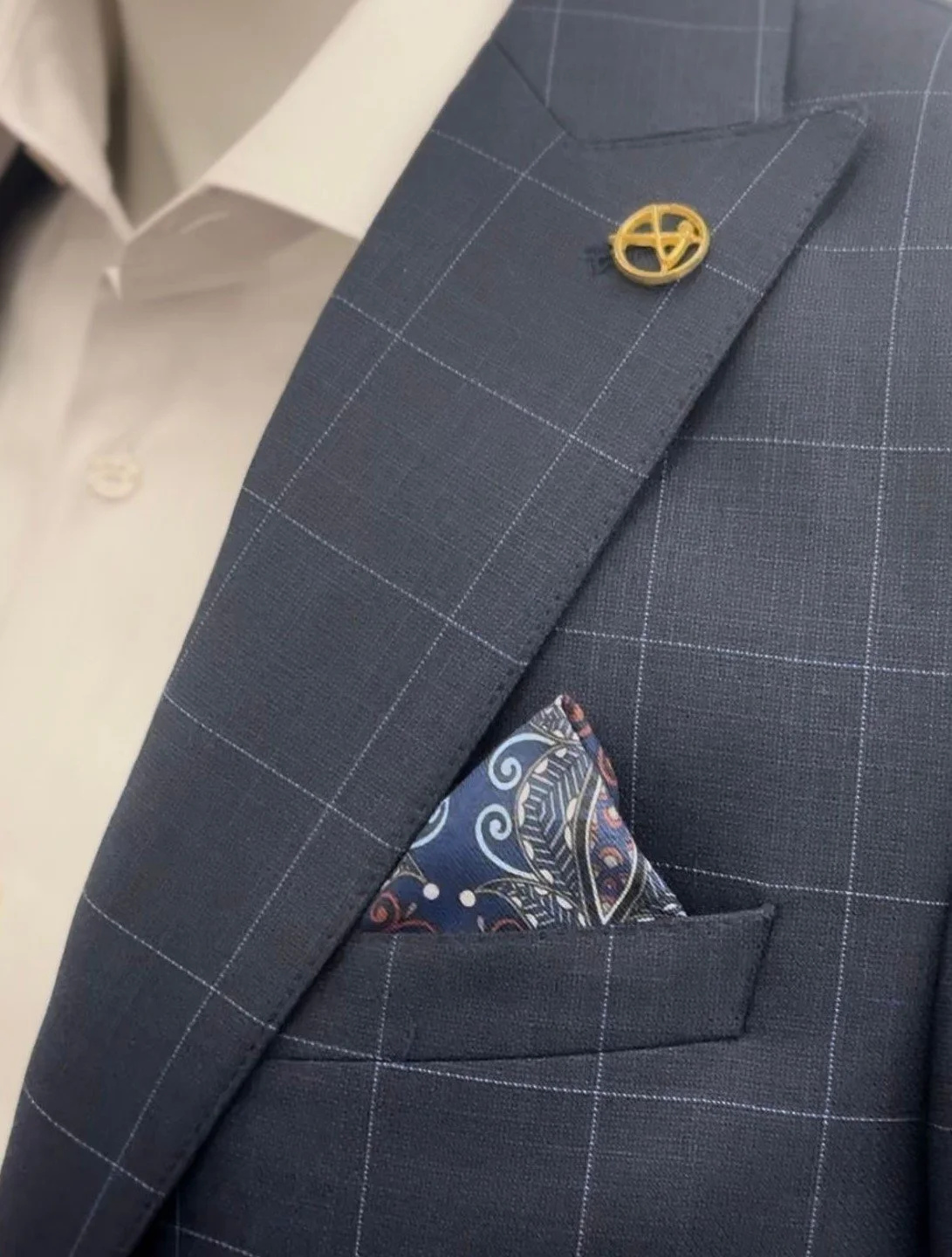 Close-up of a navy blue suit jacket with white windowpane pattern, a gold lapel pin, and a colorful pocket square in the breast pocket.