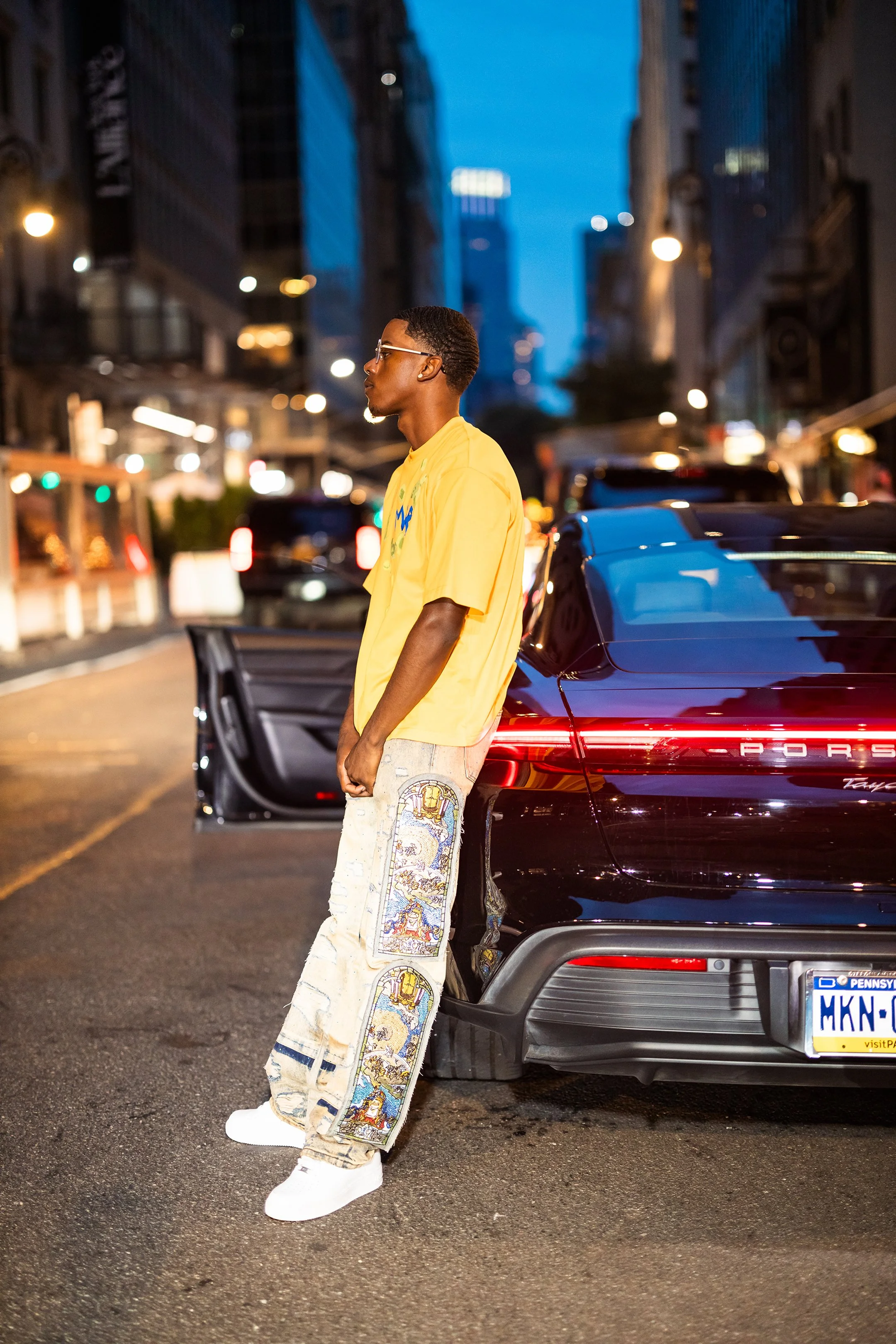 A young man wearing a yellow t-shirt, embroidered jeans, and white sneakers standing next to a black Porsche on a city street at dusk.
