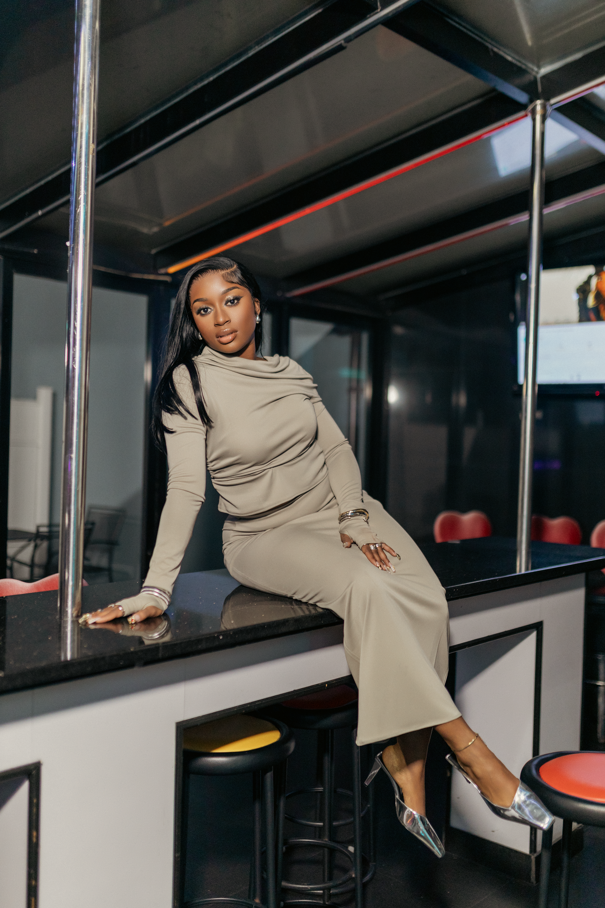A woman in a taupe dress sitting on a black counter inside a bar or club, with red and yellow bar stools, and a dark, reflective interior.