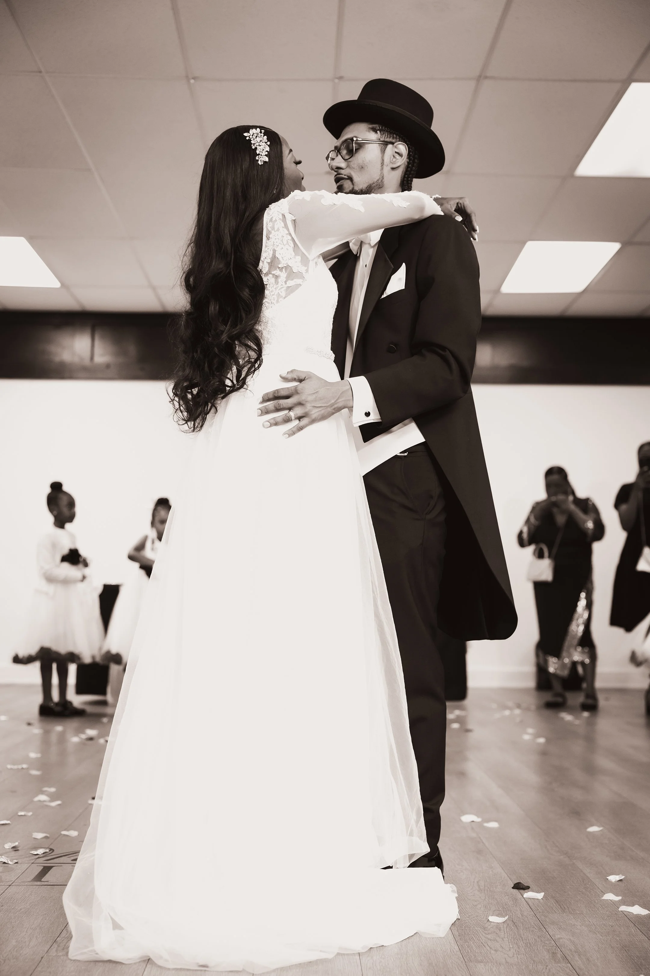 A wedding couple dancing, the bride in a white gown with long dark hair and the groom in a dark suit, white shirt, and black hat, in a reception hall with guests in the background.