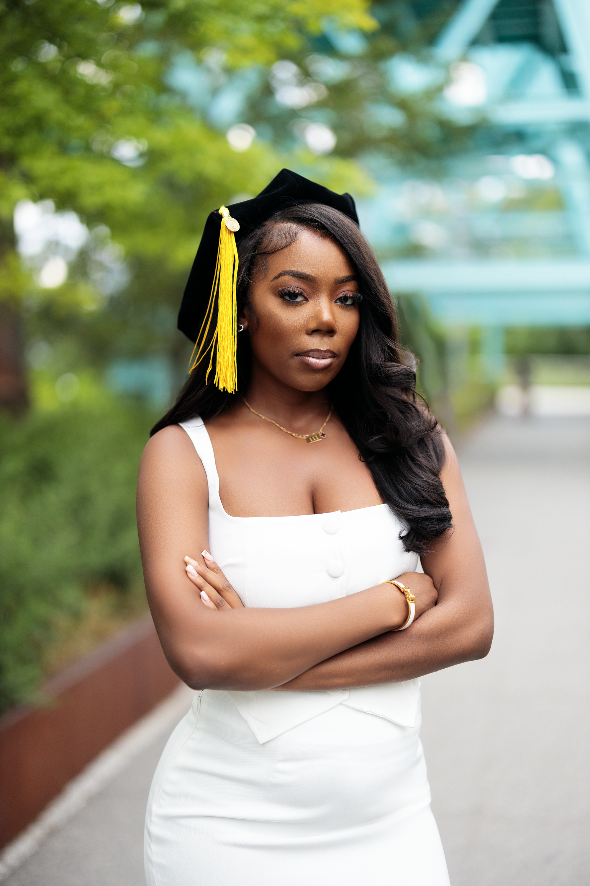 Young woman in white dress wearing a black graduation cap with yellow tassel, standing outdoors with crossed arms, green trees in the background.