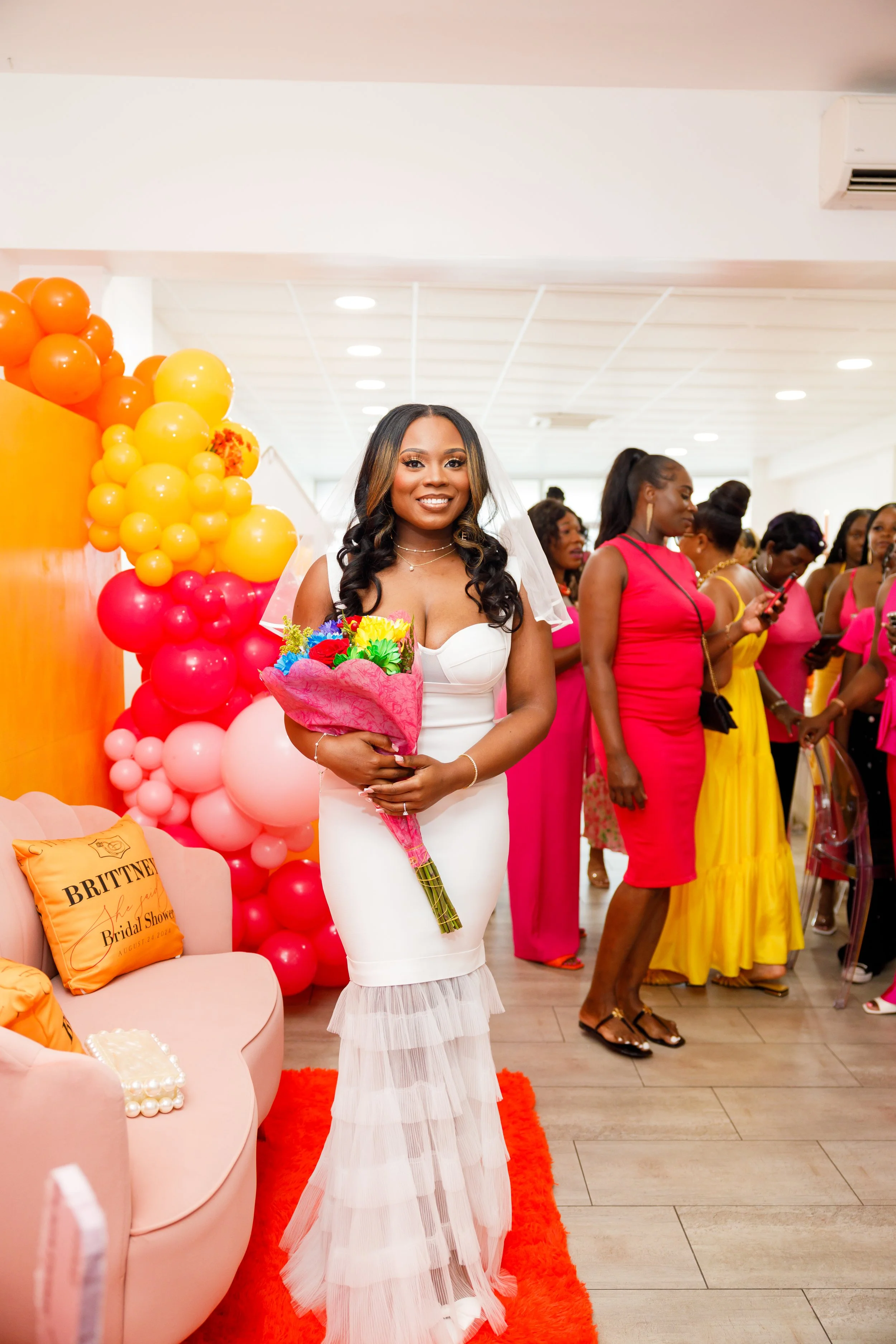 A woman in a white dress holding a bouquet of flowers at a bridal shower event, with women in colorful dresses and balloon decorations in the background.
