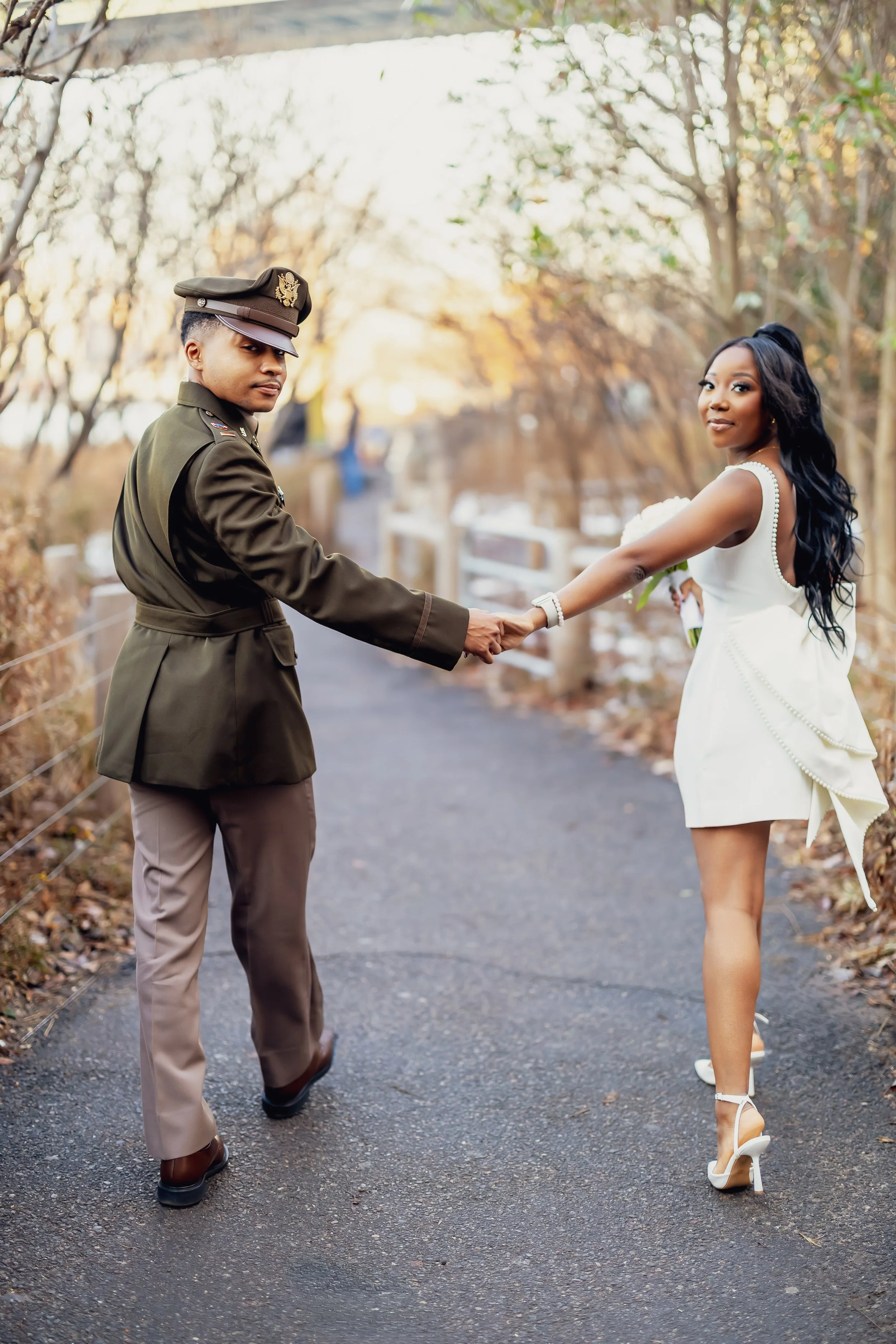 A woman in a white dress holding a bouquet, and a man in a military uniform holding hands on a wooded pathway during autumn.