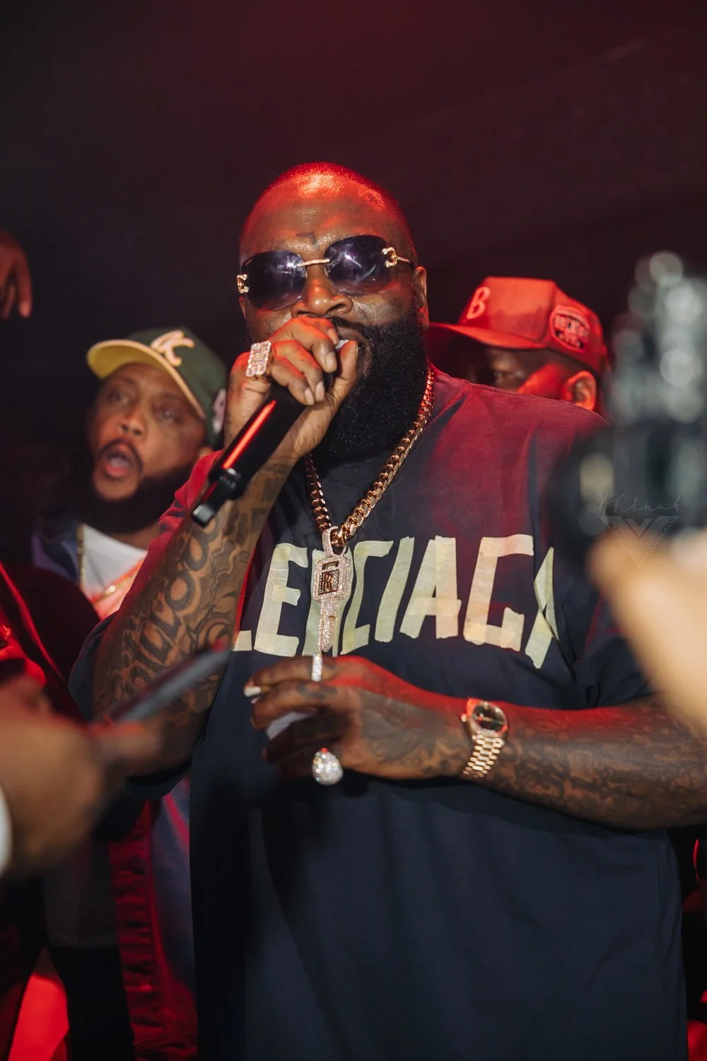 A man wearing dark sunglasses, a black T-shirt with partially visible gold text, and gold jewelry, including a large chain with a pendant, holding a microphone at a lively event with people in the background.