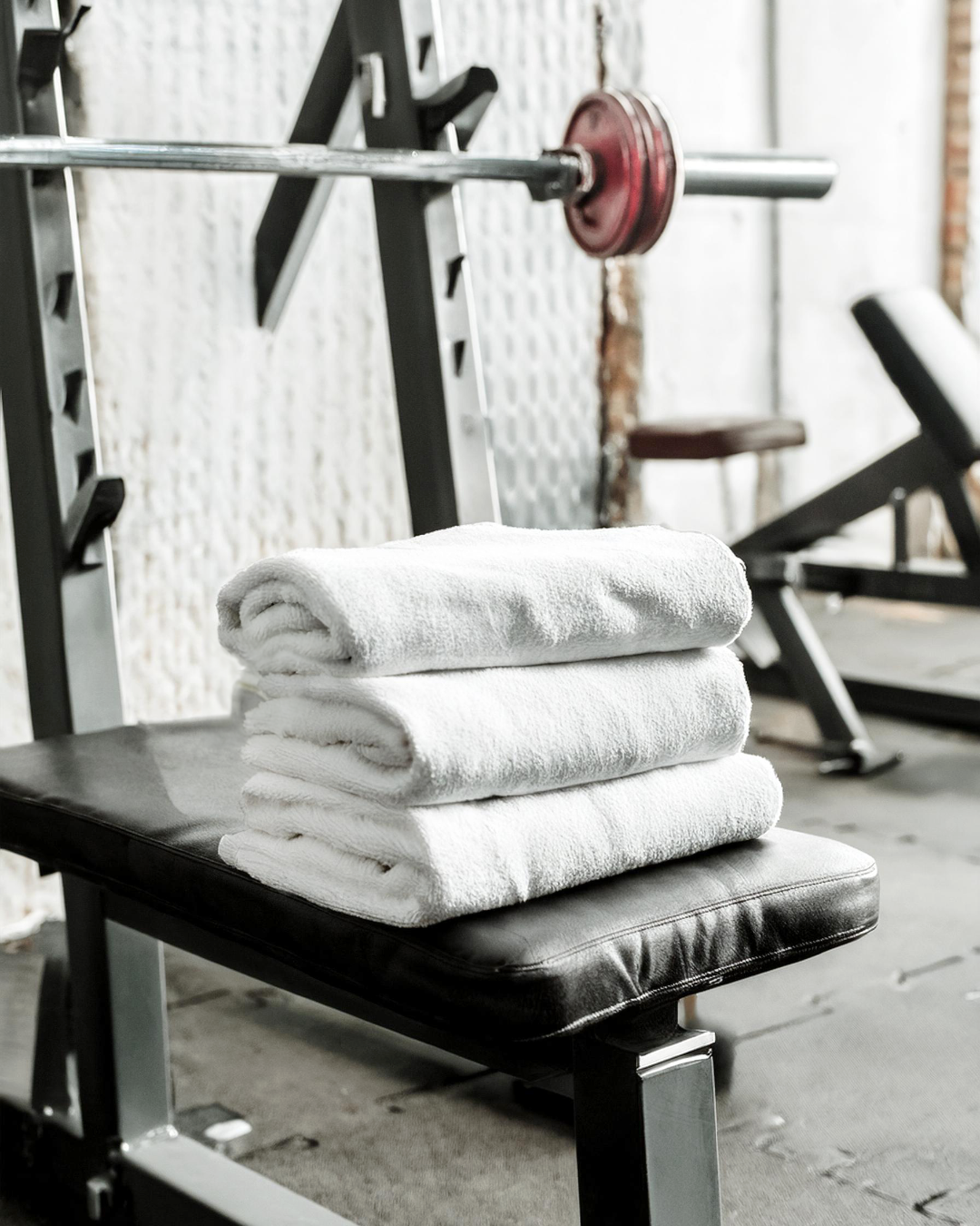 Stack of white towels on a black workout bench in a gym with weightlifting equipment in the background.