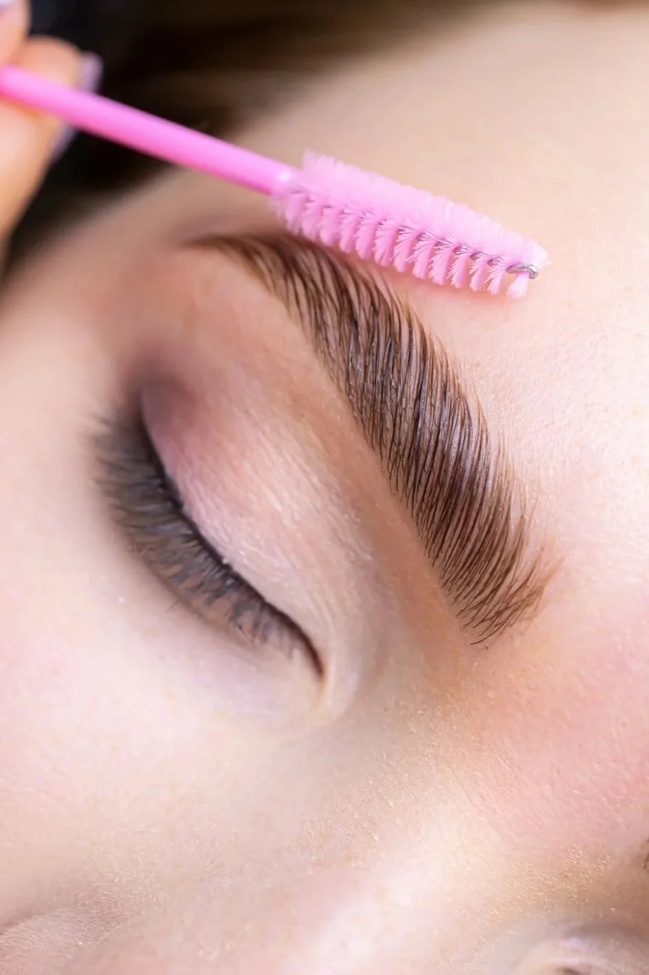 Close-up of a person's eyebrow being brushed with a pink spoolie brush.