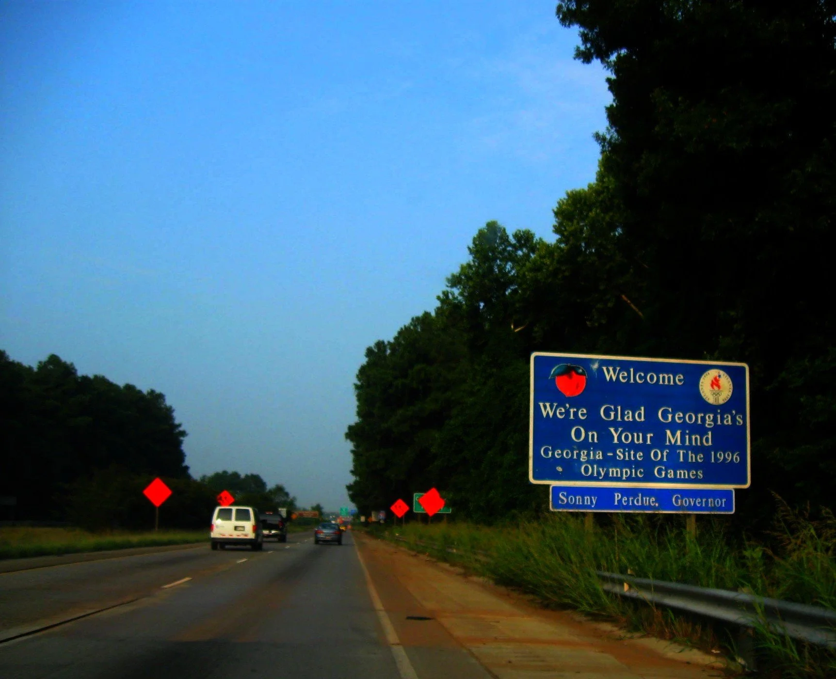 A highway with green trees on either side and a blue sign welcoming travelers to Georgia, stating it's the site of the 1996 Olympic Games with the name of Governor Sonny Perdue. Traffic is visible on the road, and the sky is clear with some clouds.