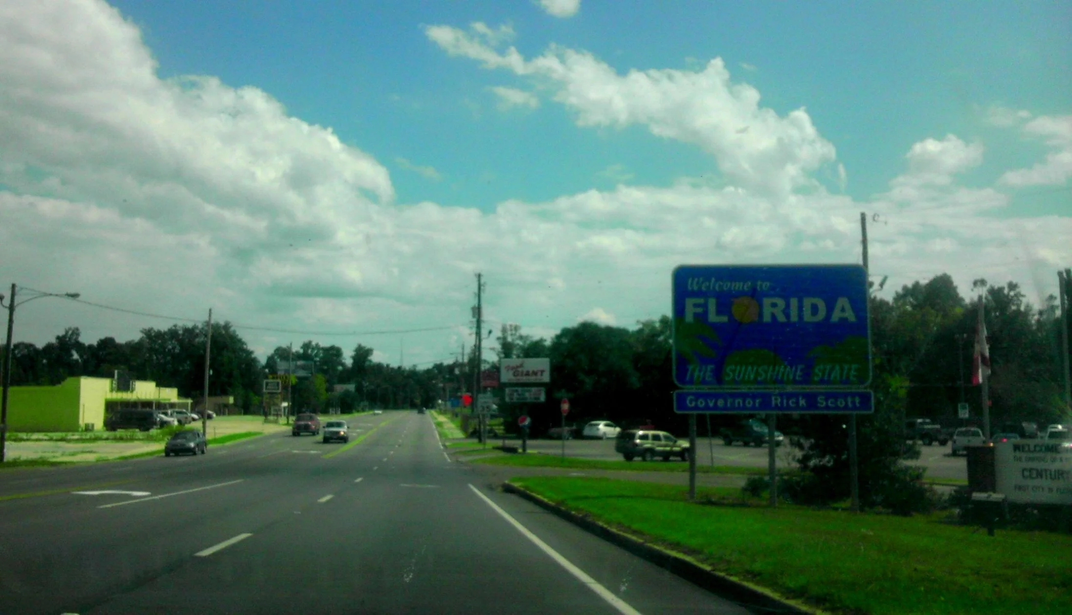 A highway in Florida with a large blue welcome sign that reads "Welcome to Florida, the Sunshine State." The sign includes the name of the governor, Rick Scott. Cars are driving on the road, and there are trees, utility poles, and billboards along th