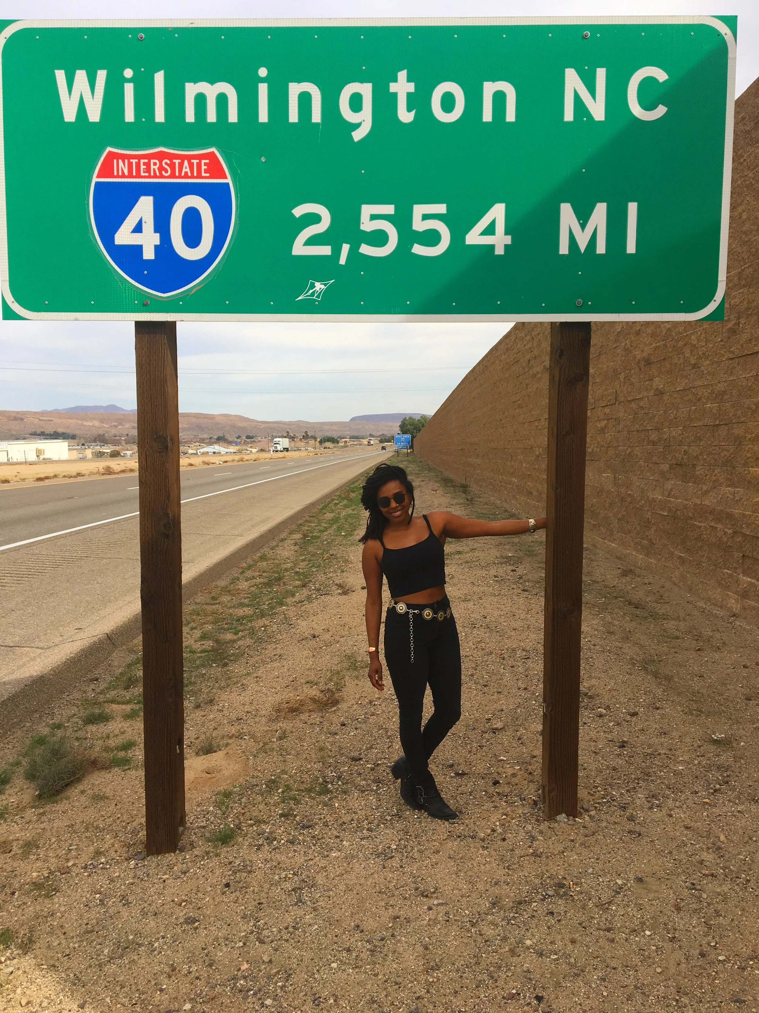Traveler posing beneath a roadside sign in Barstow, California, showing Wilmington, North Carolina, 2,554 miles away.