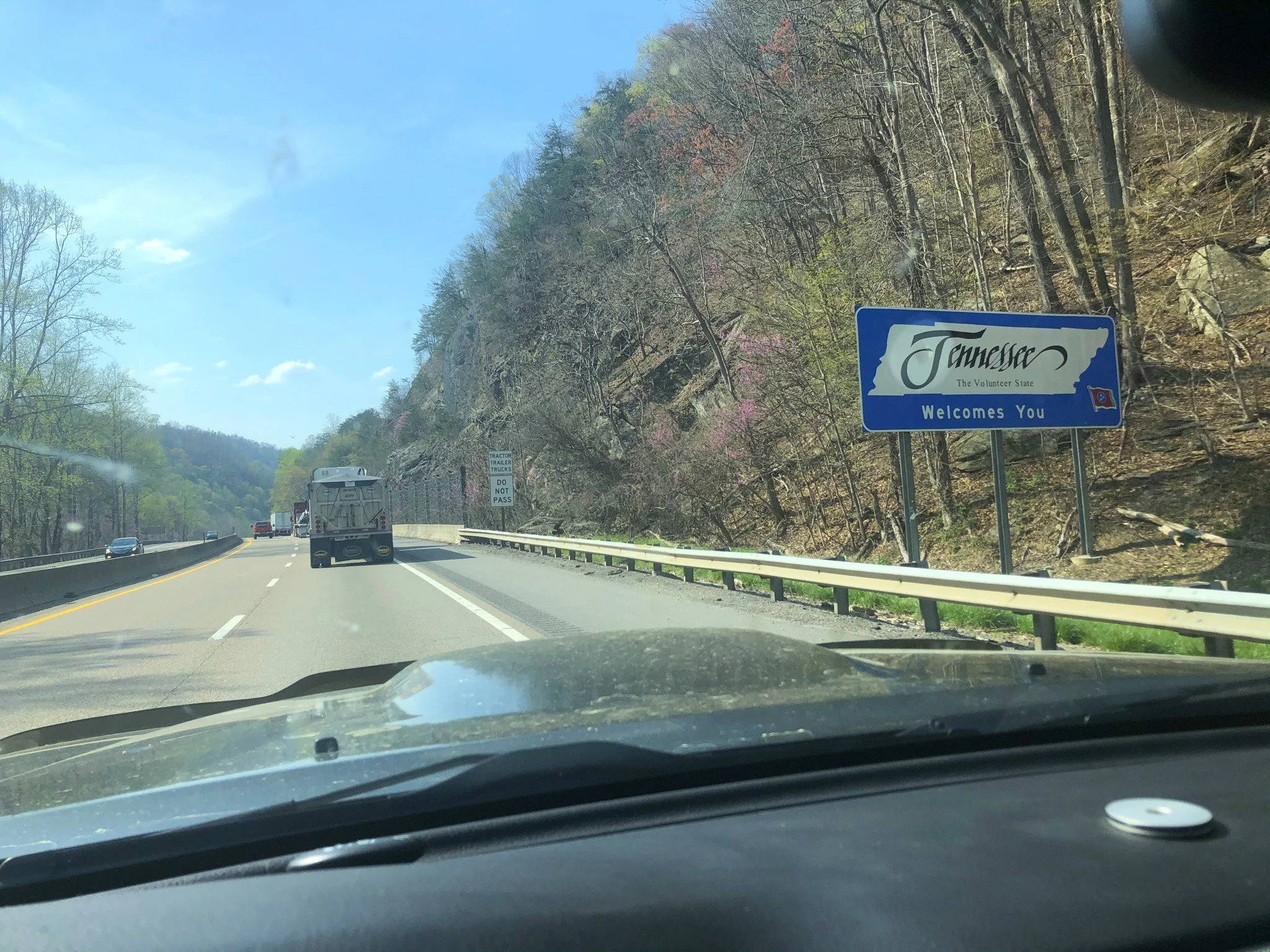 A highway with traffic and a sign that says 'Tennessee The Volunteer State Welcomes You.' The landscape includes trees on a hillside and a clear blue sky.