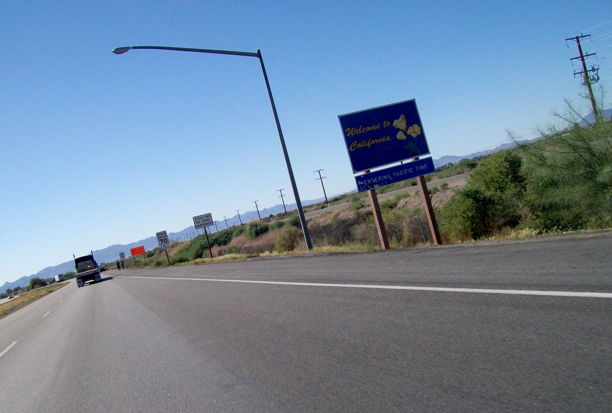 Highway scene with a blue welcome sign reading "Welcome to California" with flower illustrations, and smaller signs indicating speed limits and fines, with mountains in the background and clear blue sky.