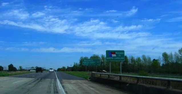 A highway scene showing a road with vehicles, green exit signs indicating directions to North Carolina and other destinations, under a bright blue sky with some clouds.